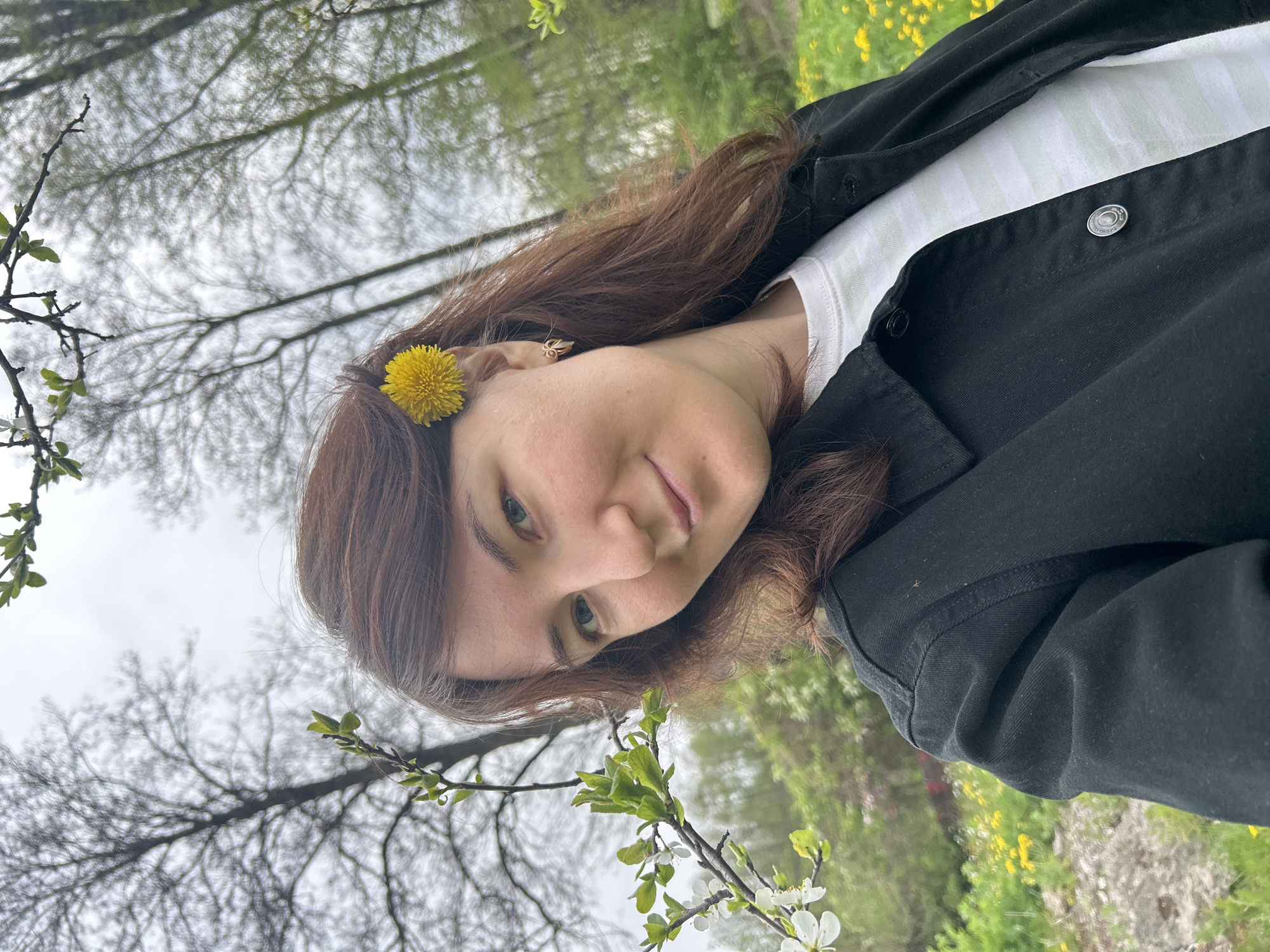 A young woman with brown hair is posing outdoors. She has a dandelion in her hair near her left ear. She is wearing a striped shirt and a black jacket. The background shows trees and greenery.
The image does not provide information about a specific location, such as the city of Noviy, Russia.
