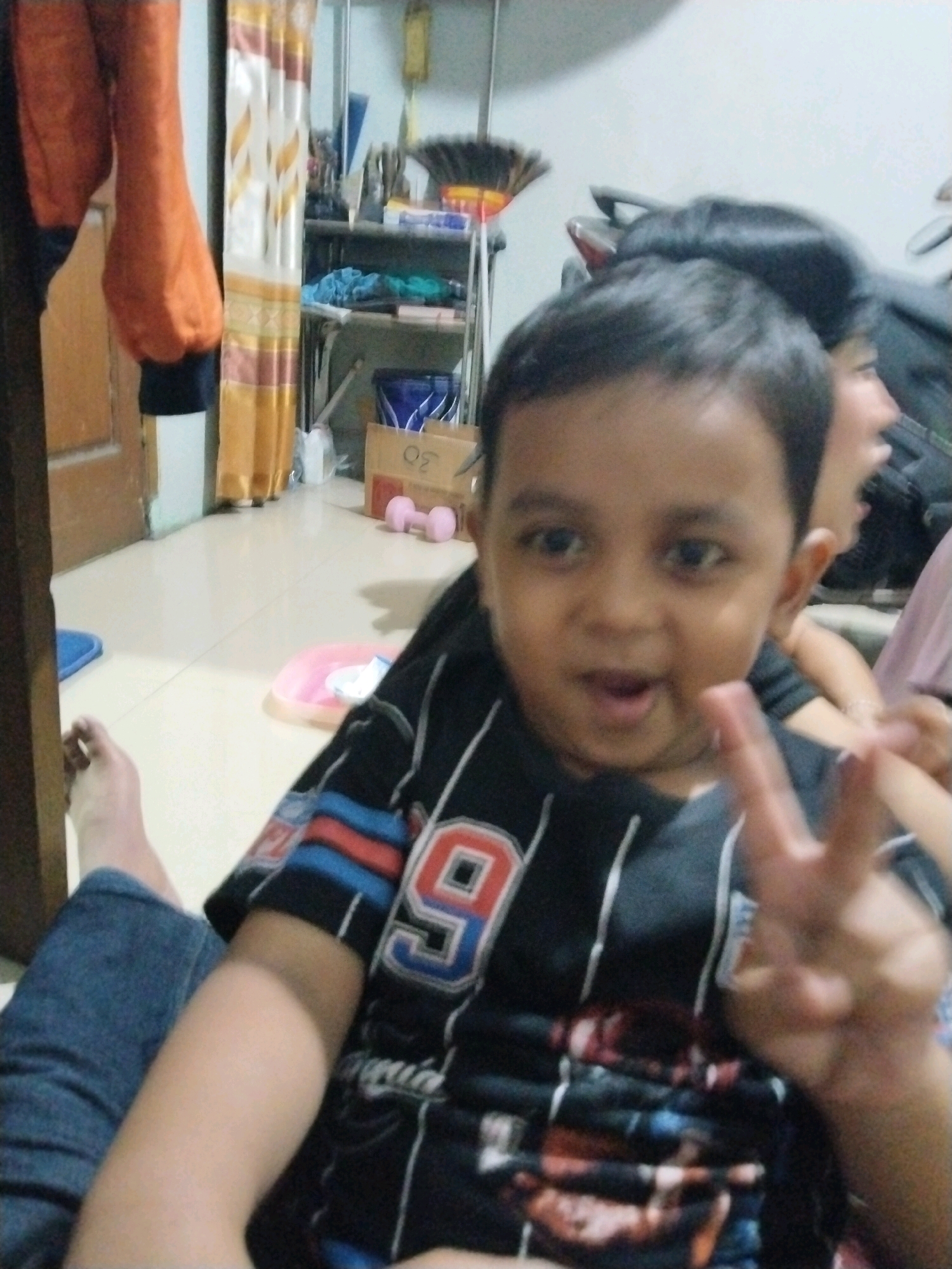 A young boy with dark hair and brown eyes is in the foreground of this indoor scene, holding up two fingers in a peace sign. He is wearing a black t-shirt with white and red stripes and a number "9" emblem. The boy is looking at the camera with his mouth open, appearing to be speaking or making a sound. Behind him, another person's head and shoulders are visible, also with dark hair. The background shows a cluttered shelf with various items, including what appears to be a broom, boxes, and blue and pink objects. The floor is tiled. The overall environment suggests a home setting.