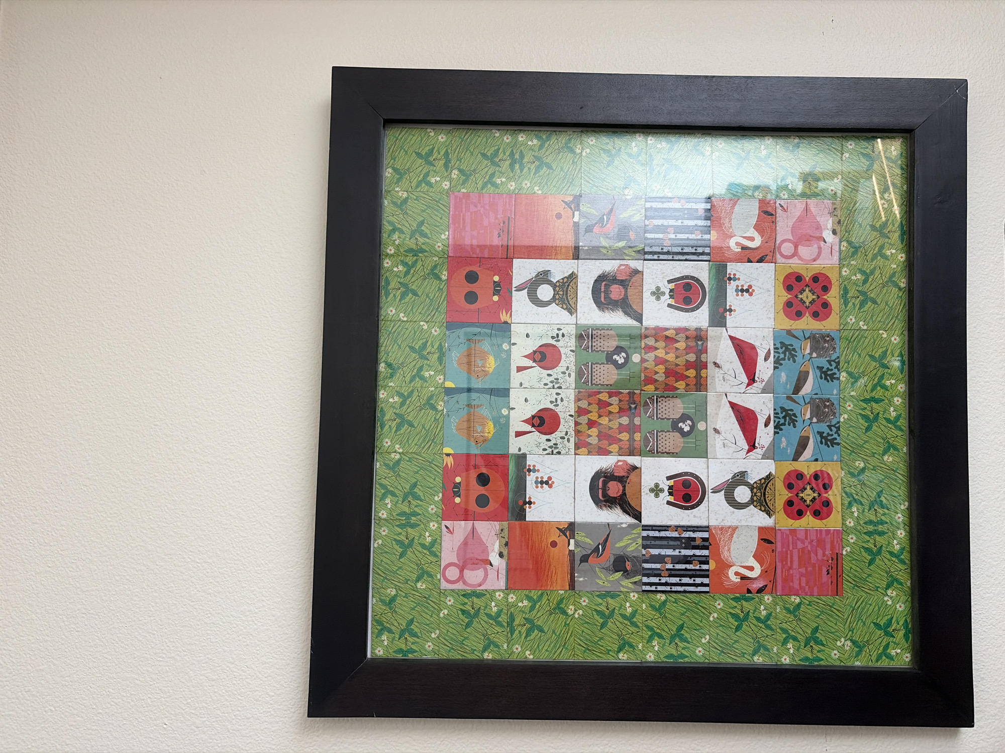 A framed piece of art is displayed on a textured white wall. The artwork itself is a grid of square images, each depicting a different illustration, set against a green background with a repeating pattern of leaves and small white flowers. The grid appears to be 6 squares wide by 6 squares tall, though the outer edges of the artwork are partially obscured by the black frame.
The individual squares within the grid feature a variety of colorful and whimsical designs. Some squares contain illustrations of animals, such as flamingos, birds, a monkey, and a swan. Others depict insects like ladybugs and possibly other creatures or stylized patterns. There are also squares with abstract geometric designs and what appear to be representations of faces or masks. The art style is varied, with some illustrations having a more graphic, modern feel, while others might be inspired by folk art or children's book illustrations. The colors are vibrant and diverse, ranging from bright reds, oranges, and yellows to blues, greens, and pinks.
The frame is a simple, black, rectangular border that surrounds the artwork. The wall behind the artwork is a neutral, off-white color with a slightly textured finish. The lighting in the scene appears to be ambient, casting a soft illumination on the artwork and wall. There are no people, actions, or interactions visible in the image. The scene is static and focuses solely on the artwork and its presentation on the wall.
This image does not provide any information about the specific location within the city of Vancouver, United States of America.