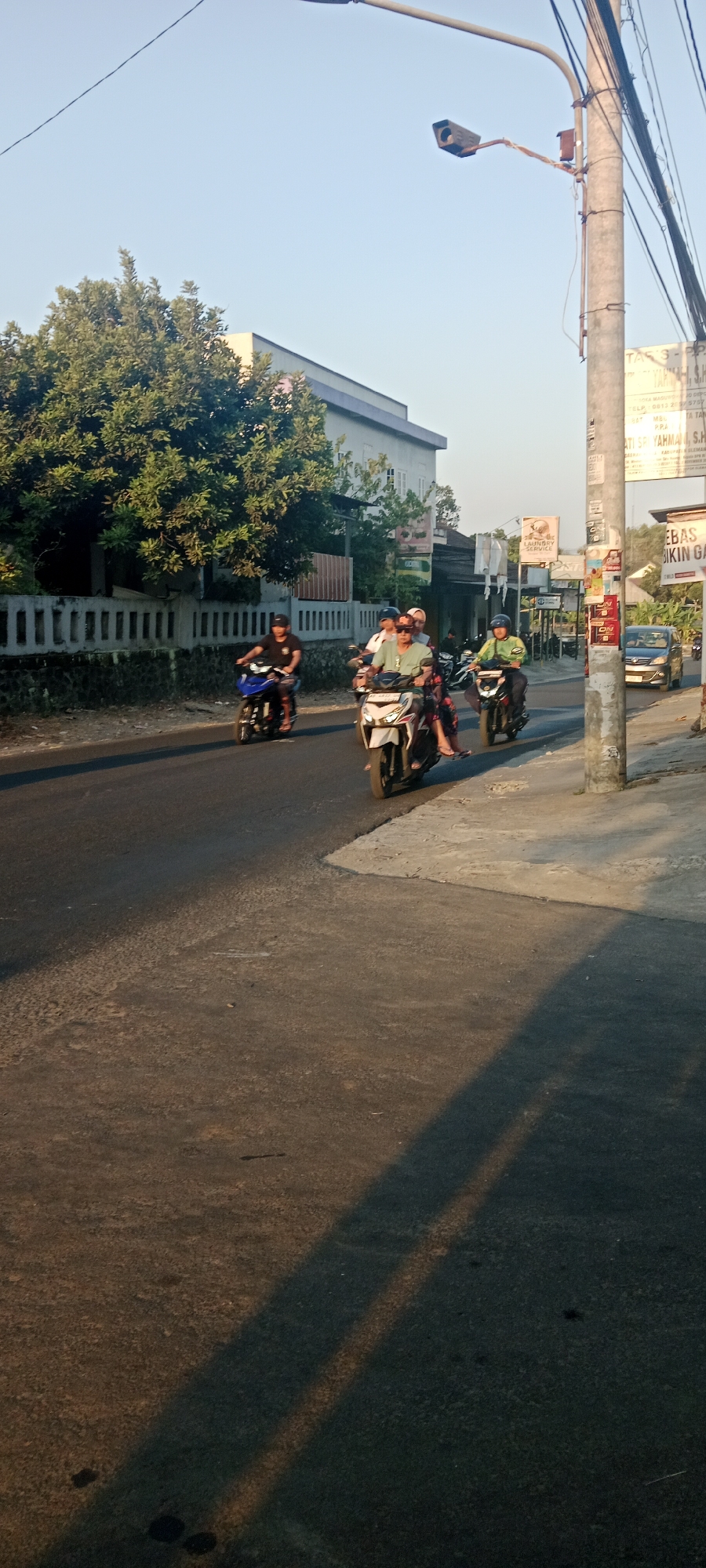 The image depicts a street scene in Maguwoharjo, Indonesia, at what appears to be late afternoon or early evening, given the long shadows cast across the road. The perspective is from a slightly elevated position, looking down a paved road lined with trees and buildings on the left, and a sidewalk and utility pole on the right.
Several people are riding motorcycles, moving from left to right. In the foreground on the left, a person wearing a black shirt and a black cap is riding a blue and black motorcycle. Next to them, slightly ahead, is a white and black motorcycle with two occupants; the rider wears a light green shirt and a helmet, and the passenger, likely female, wears a patterned dress and a headscarf. Further to the right, a person in a light green shirt and helmet is riding a dark motorcycle. In the background, another motorcycle is visible, and a dark-colored car is parked on the right side of the road.
The environment includes lush green trees and a building with a light-colored facade and a blue trim. A white fence with a dark stone base separates the road from the property on the left. On the right, a concrete sidewalk is present, and a tall utility pole with various wires and attached equipment, including what appears to be a camera or sensor, stands prominently. Several signs are attached to the utility pole, with some text legible, including "TAS-PP" and an advertisement for "LAUNDRY KLINIK." The road surface is asphalt, with some visible patches and wear. The sky is clear and a pale blue.