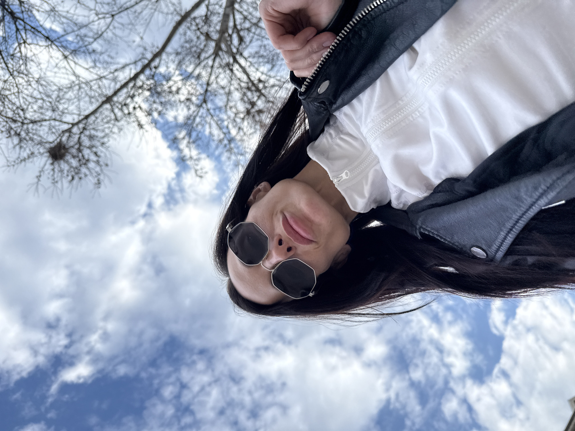 The image presents a low-angle, upward-looking view of a single female individual outdoors. Her face, positioned centrally, is largely obscured by dark, octagonal-shaped sunglasses with silver frames. She has dark, long hair extending over her shoulders. Her attire includes a black leather jacket featuring a prominent metal zipper and a snap button on the lapel, worn over a white, collared garment with a white zipper. The individual's right hand is visible, gripping the zipper pull of the black jacket. The background consists of a blue sky partially covered by white clouds. Bare tree branches, characteristic of deciduous trees in a dormant state, are visible in the upper right and right side of the frame. A bird's nest is discernible within the bare branches in the upper right. The environment suggests an outdoor setting, potentially a park or urban green space. No specific architectural or geographical landmarks are present to definitively identify a precise location within the city of Pleven, Bulgaria.