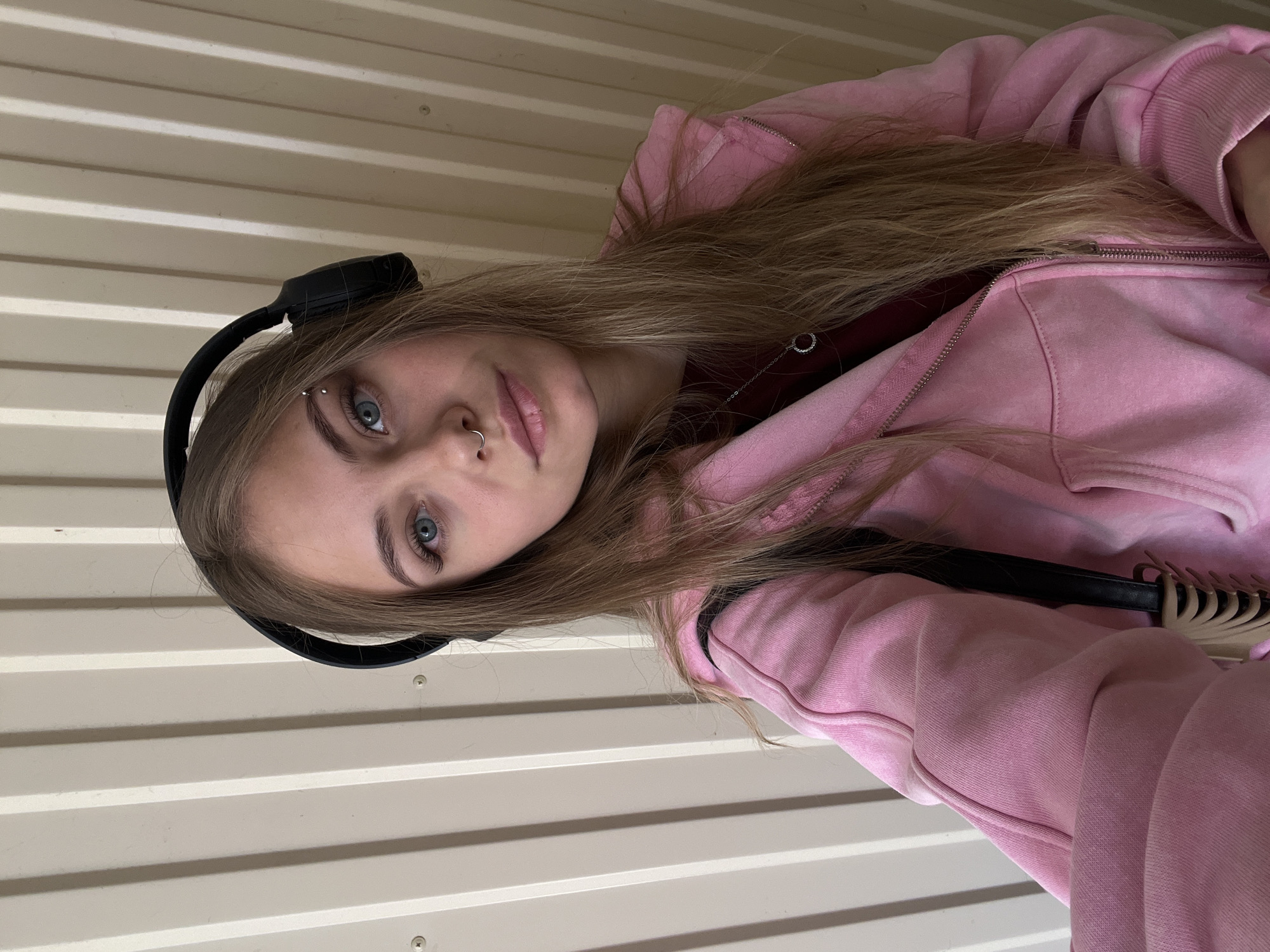 A selfie features a young person, likely female, with long, light brown hair, fair skin, and striking blue eyes. They are wearing black over-ear headphones and a pink zip-up hoodie, partially unzipped to reveal a darker red or maroon top underneath. Notable facial piercings include a septum ring and an eyebrow barbell on their left brow. The person holds a neutral expression while looking directly at the camera. A black strap, possibly from a backpack or bag, is visible over their right shoulder. The background consists of a light-colored, horizontal corrugated metal or siding wall with visible screws, suggesting an outdoor or semi-sheltered urban/industrial setting. The lighting appears natural, indicating the photograph was taken during the daytime. No visible text is present in the image. The location context is Chaikovsky, Russia.