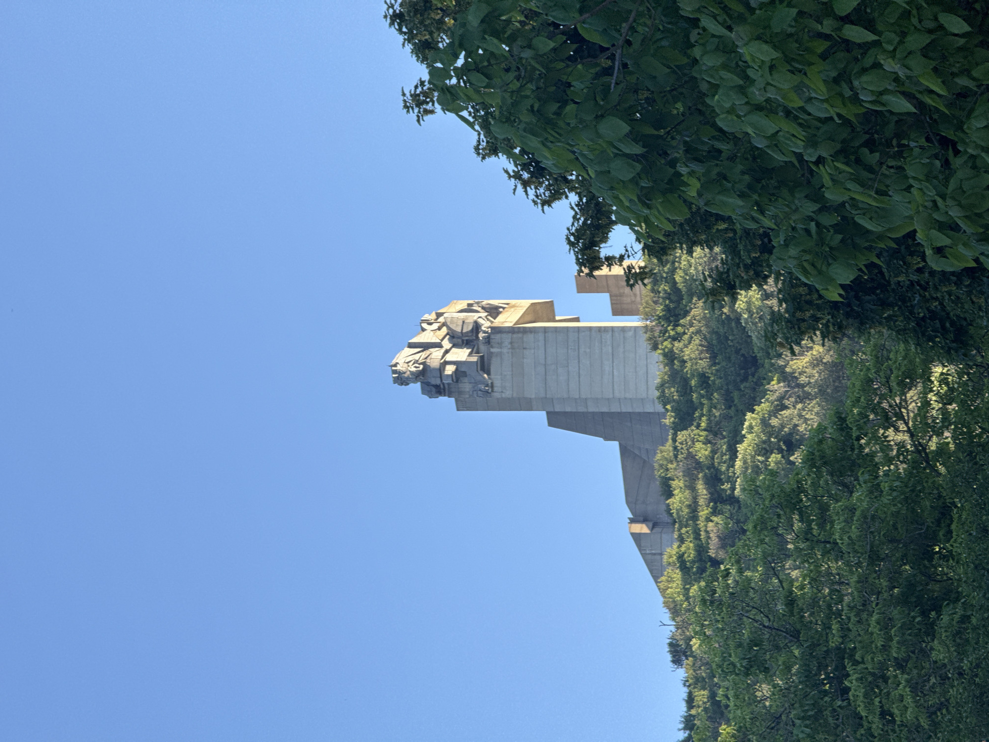 The image shows the Founders of the Bulgarian State Monument in Shumen, Bulgaria, set against a clear blue sky. The monument, a large concrete structure with a sculpted frieze at the top, is partially framed by green trees and vegetation on the right side of the image. The perspective is a low angle, emphasizing the monument's height.