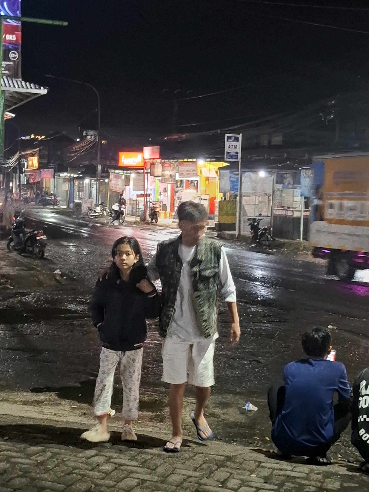 In the city of Ciheulang Tonggoh, Indonesia, at night, two individuals, a young female and a young male, stand close together on a wet, paved street. The young female, on the left, wears a dark jacket and patterned pants. The young male, on the right, wears a white shirt and a camouflage vest with white shorts. Behind them, a street is lined with shops and stalls, illuminated by various lights. Several motorcycles are parked along the street. In the foreground, on the right side of the image, a person wearing a blue t-shirt is crouched down looking at a red mobile phone. The ground is wet and reflective, indicating recent rain. A large truck is partially visible on the right side of the frame.