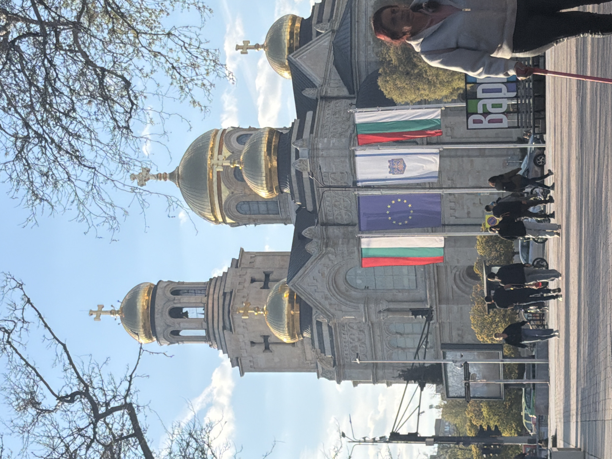 The image depicts a daytime street scene in Varna, Bulgaria, featuring the Dormition of the Theotokos Cathedral. In the foreground, bare tree branches frame the top left. The main subject is the grand cathedral, an ornate Eastern Orthodox building with multiple golden domes, each topped with a cross. Its stone facade is intricately detailed.
On the right side of the image, facing the cathedral, is a paved square or street where several people are present. One woman in the immediate foreground, wearing a light-colored hoodie and dark pants, holds a red stick-like object (possibly a selfie stick or a cane) and looks towards the camera. Further back, other pedestrians are seen walking or standing.
Several flags are displayed vertically on poles to the right of the cathedral. From top to bottom, they include the Bulgarian flag (white, green, red horizontal stripes), a blue flag with a distinct emblem (likely Varna's municipal flag), the European Union flag (blue with yellow stars), and another Bulgarian flag.
In the background, behind the people and flags, a large, colorful sign is partially visible, displaying the Cyrillic text "Варна" (Varna), confirming the location. A car is also partially seen. The sky is bright and clear with scattered clouds, indicating good weather. The overall scene captures daily life in front of a prominent landmark.