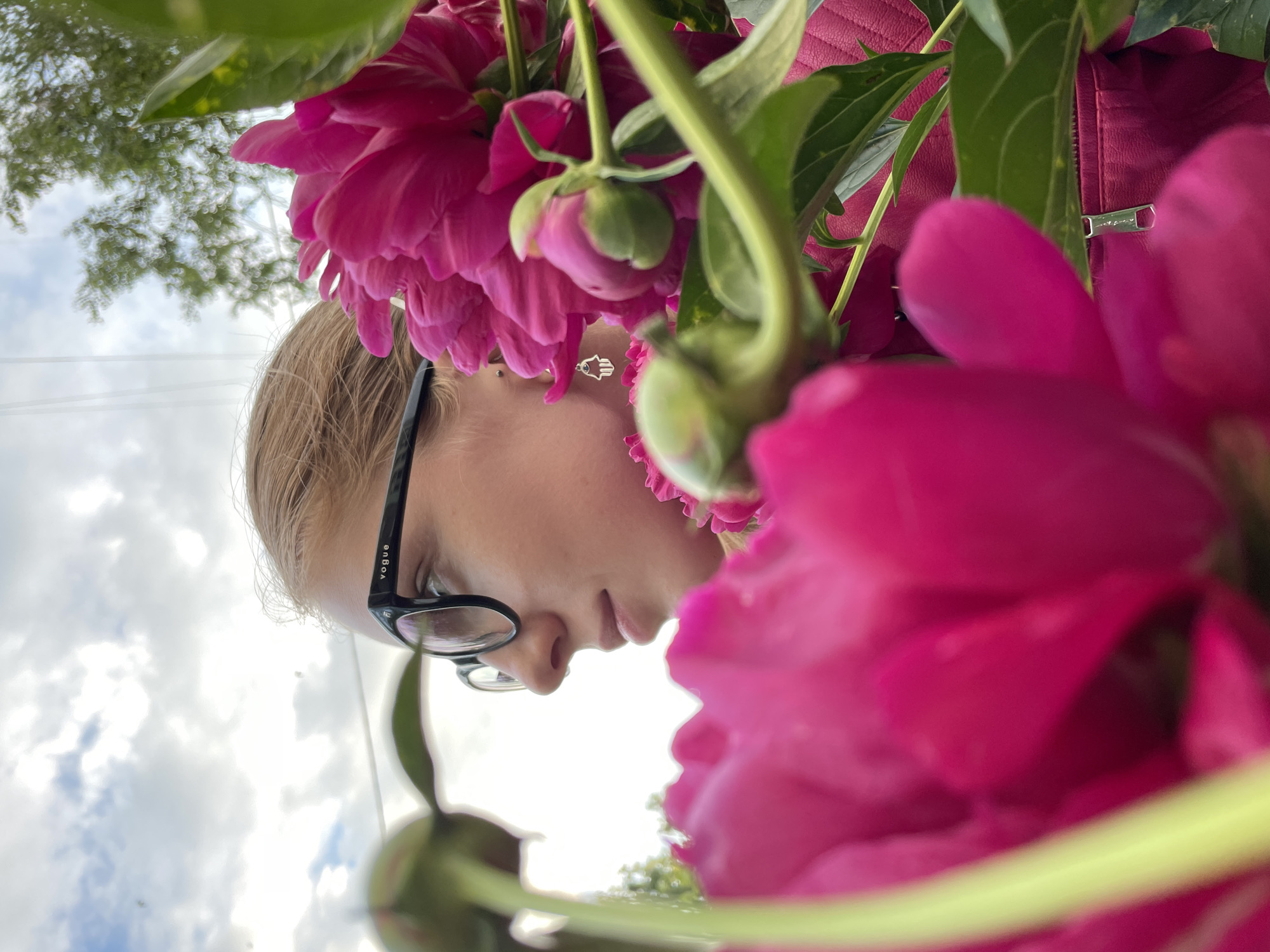 A young woman wearing black-rimmed glasses with "Vogue" inscribed on the arm is partially obscured by vibrant pink peonies. Her hair is light brown and pulled back. She is looking off to the left, with a neutral expression. A silver earring shaped like a hamsa hand is visible. She is wearing a pink jacket with a zippered pocket. The background shows a cloudy sky and green foliage. The scene is outdoors, likely in a park or garden setting within Izhevsk, Russia.