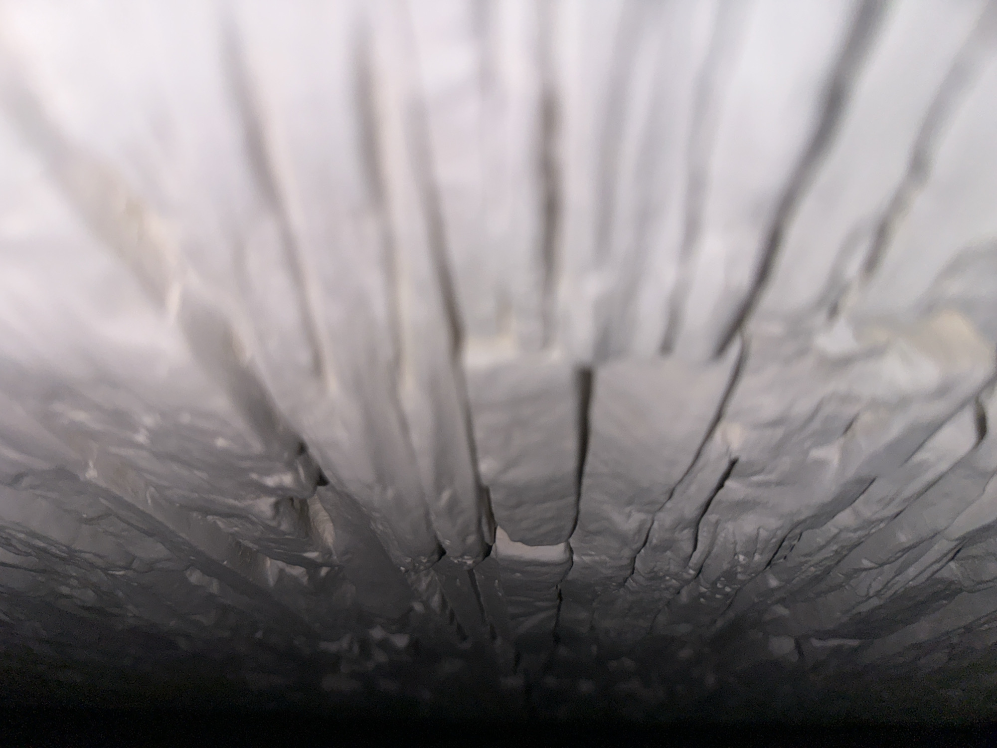 The image presents an abstract, close-up view from a low-angle perspective, looking upwards into what appears to be a textured, cavern-like space. The primary subject is a surface characterized by numerous vertical folds and creases, predominantly in shades of silvery-grey and white.
The scene gives the impression of being inside a confined area, possibly a container, package, or a vent. The top portion of the image is significantly brighter and more out-of-focus, suggesting a light source or an opening, while the bottom transitions into complete darkness, implying depth or an unlit recess.
The texture of the material appears somewhat reflective and crinkled, resembling aluminum foil, a metallic insulation material, or a heavily textured fabric. The folds create a strong sense of verticality and depth, drawing the eye upwards. There are no discernible people, objects other than the textured surface itself, or any specific activities taking place.
Notable details include the shallow depth of field, with the sharpest focus on the middle ground of the folded material. The lighting appears diffuse and artificial, or like bright ambient light from above. There are no clear location cues beyond the structural characteristics of the material itself. No visible text is present in the image. The overall impression is one of an unusual, abstract pattern created by light, shadow, and crumpled surfaces.