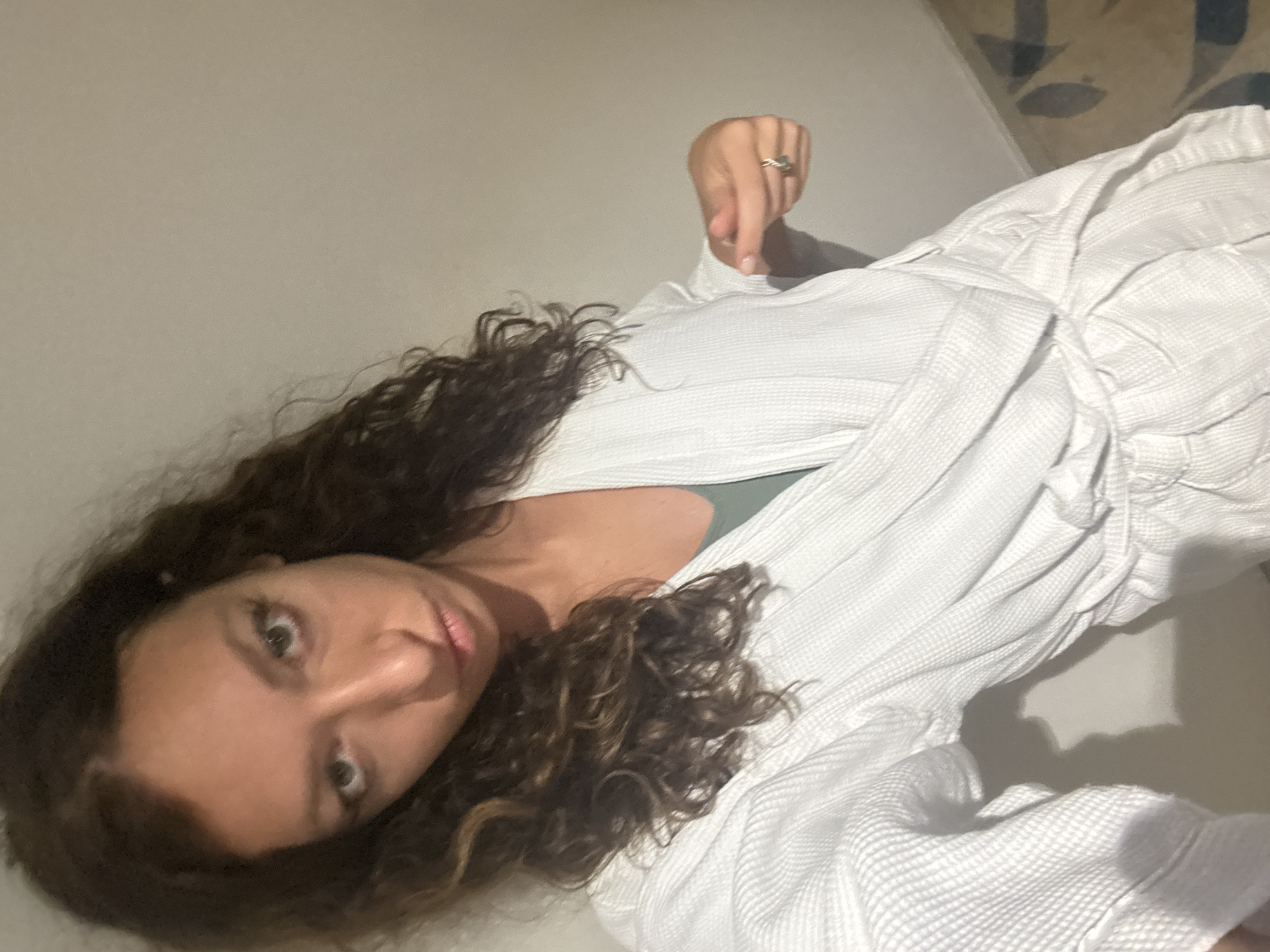 A woman with long, curly brown hair is lying on a white surface, possibly a bed. She is wearing a white, waffle-patterned robe. Her head is tilted towards the camera, and she is looking directly at the viewer. She is wearing a ring on her right hand, which is slightly raised. The background is a plain white wall. The scene appears to be indoors and has soft lighting. The image does not contain sufficient detail to determine the location in Đà Nẵng, Vietnam.
