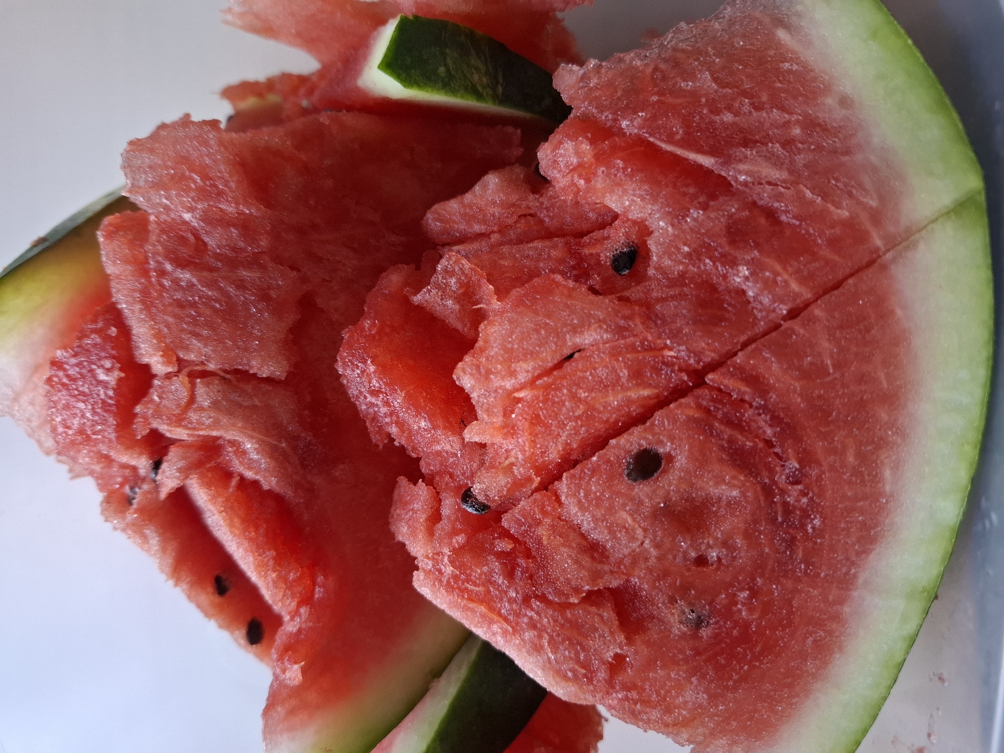 A close-up, overhead shot features a pile of freshly cut watermelon slices. The vibrant red flesh, speckled with black seeds, contrasts with the green rind. Some slices are jagged and broken, suggesting they were recently prepared or handled. The background is a plain, light color, drawing all attention to the juicy fruit. This image captures a refreshing snack, likely prepared at home and not associated with any specific location in Tyumen, Russia.
