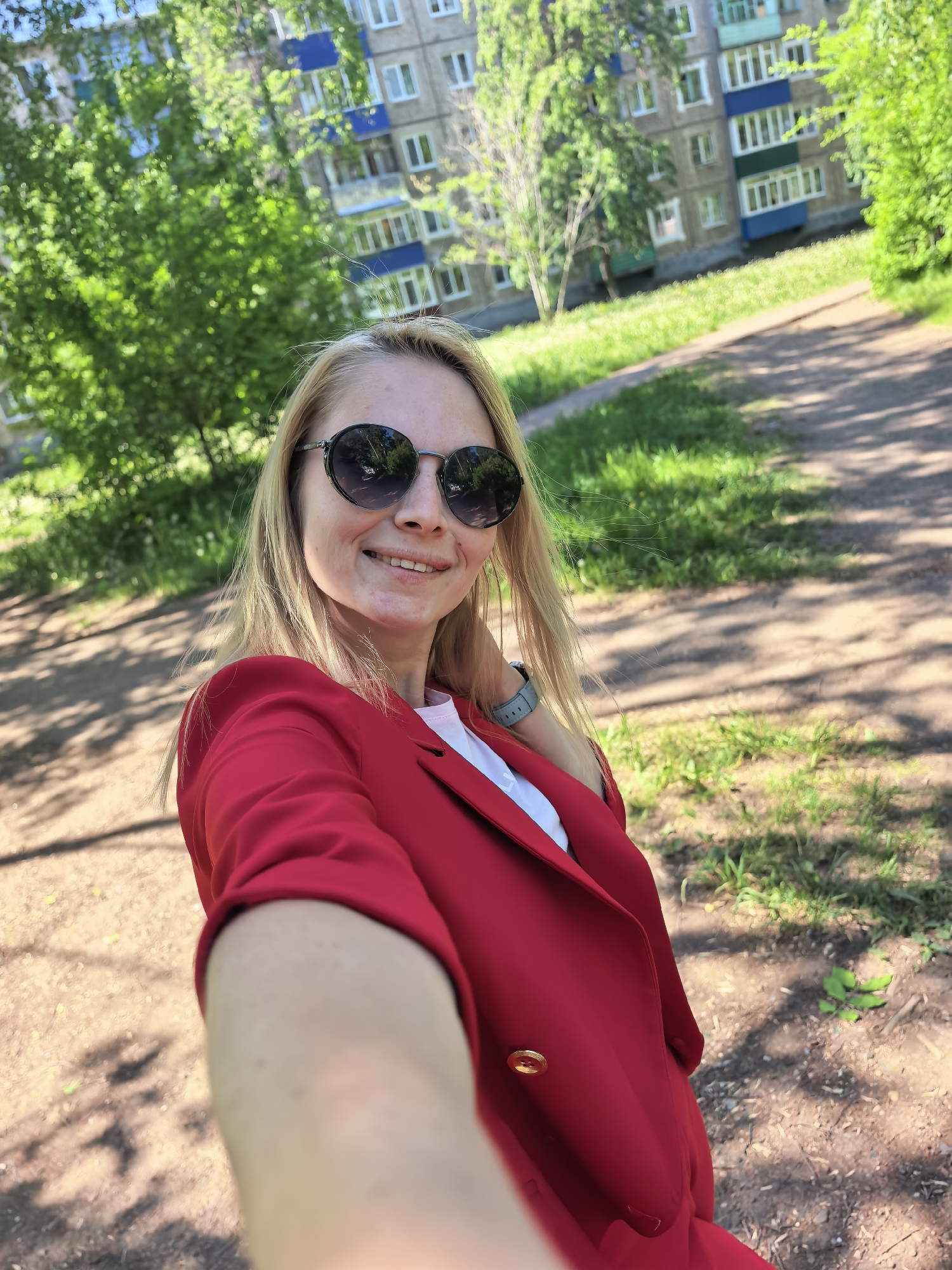 A woman is taking a selfie outdoors. She has blond hair and is wearing round sunglasses and a red blazer over a white top. A watch is visible on her left wrist. The background shows a city park setting with green trees and grass. In the distance, an apartment building is visible. The location is identified as Sterlitamak, Russia. The time of day appears to be daytime, indicated by the lighting and shadows.