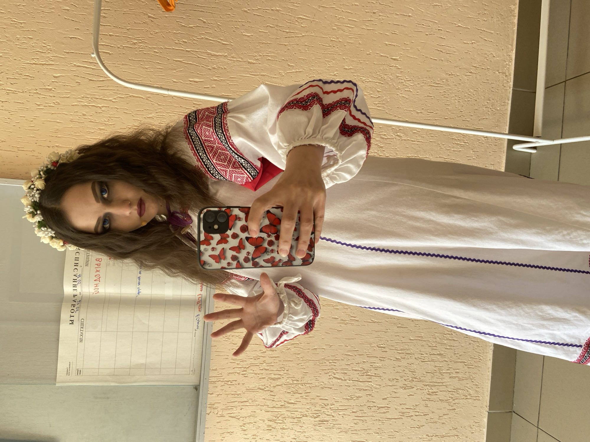 A person is posing indoors, holding a phone with a butterfly-themed case, wearing a white dress with red and purple embroidery and a floral headpiece, next to a wall with a document posted on it. The location is in Chaikovsky, Russia.