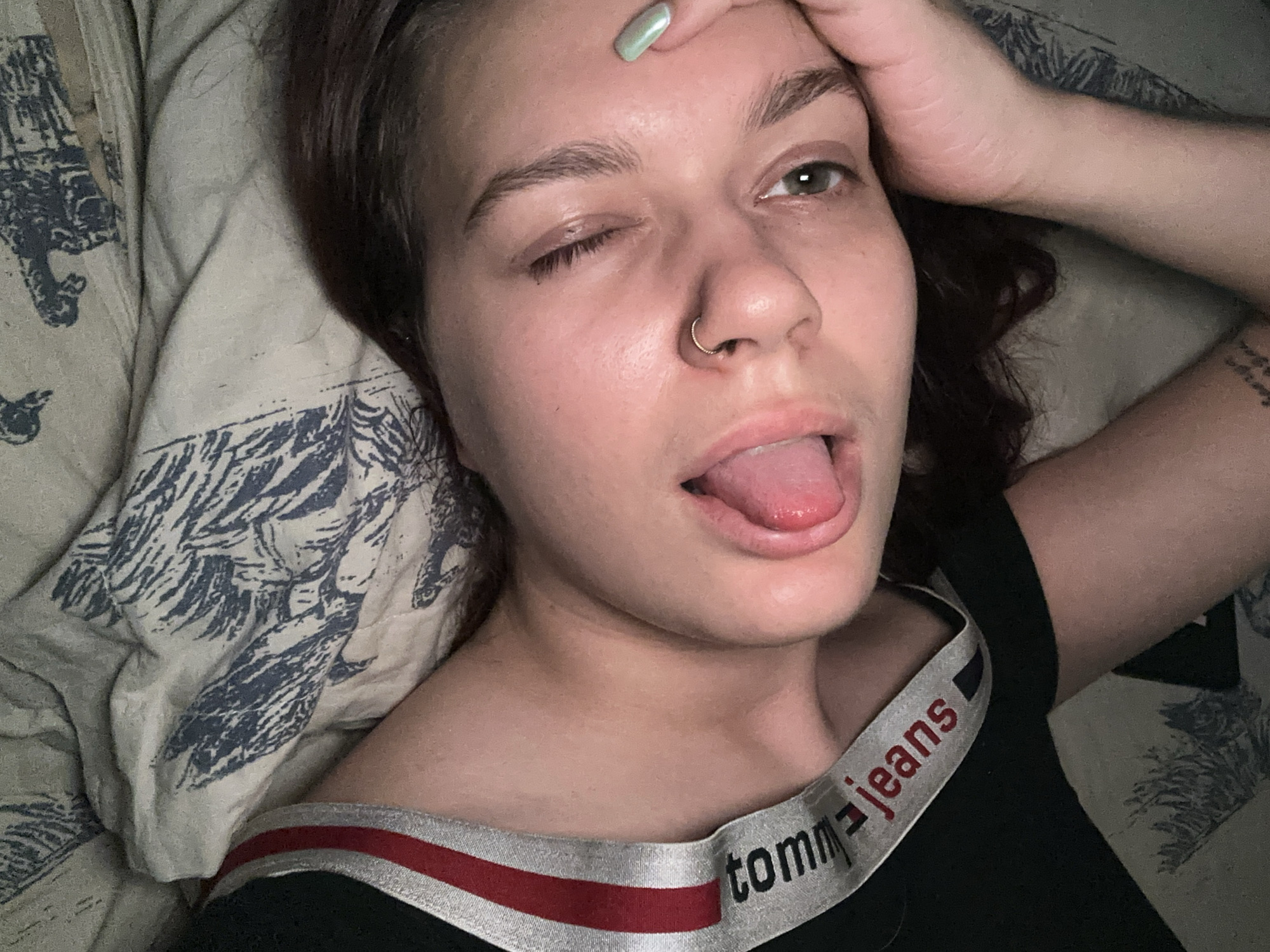 The image shows a close-up, overhead view of a young person lying down. The person has dark hair, a septum piercing, and appears to be winking with their tongue slightly out. They are wearing a black garment with a red and white striped band that says "tommy jeans". The background appears to be a bed with a patterned comforter. There are no identifiable landmarks or contextual clues that would place this image in Bishkek, Kyrgyzstan.
