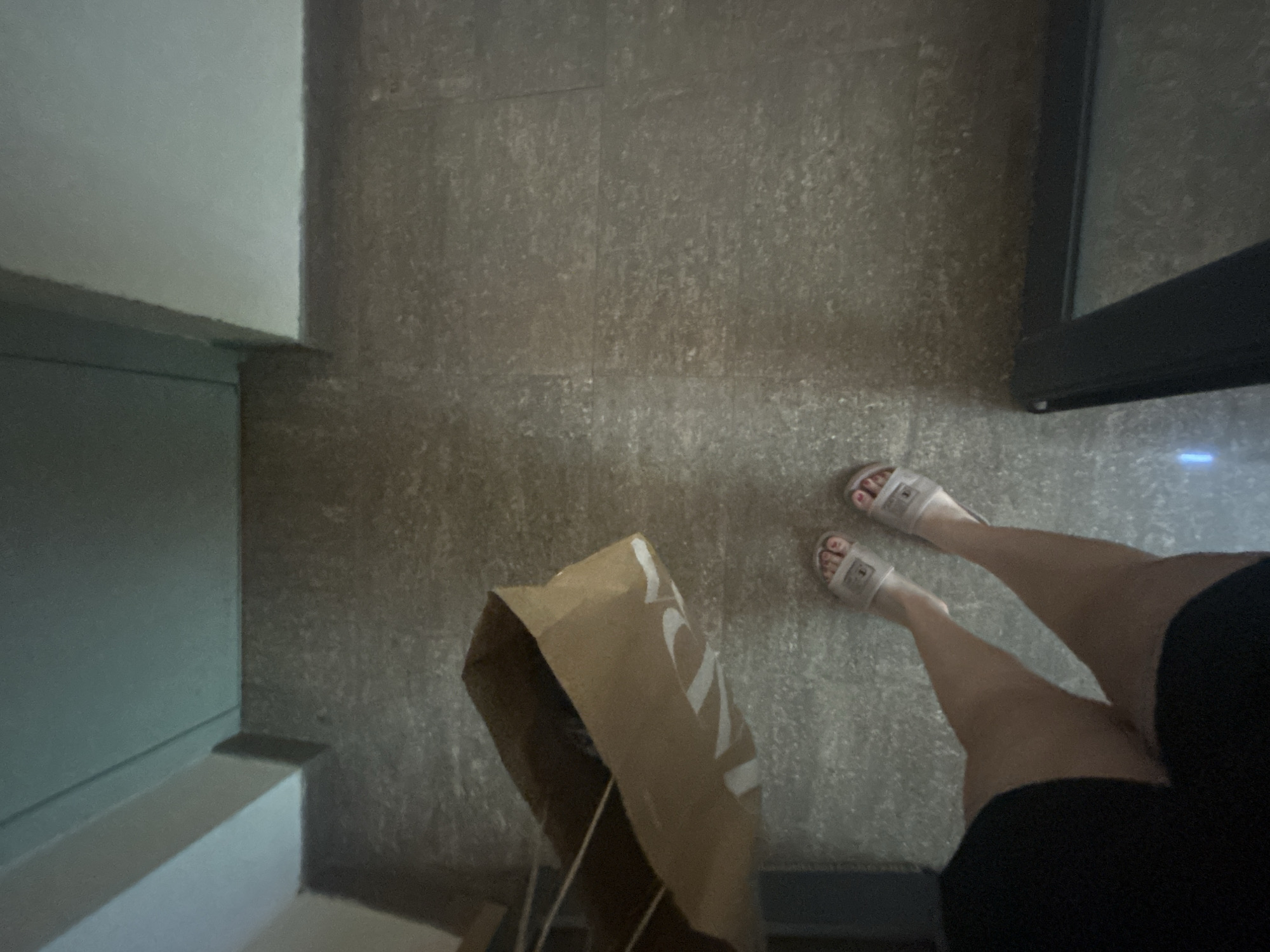 The media file shows a top-down view of a person's legs and feet, wearing white slides with a logo, standing on a speckled tiled floor. A brown paper shopping bag with a white logo is placed on the floor in front of them. The setting appears to be indoors, possibly a hallway or an entryway, with a glimpse of a pale green wall and a darker area that could be a doorway or a piece of furniture. The lighting is dim, suggesting it could be evening or a dimly lit indoor space. The person's toenails are painted pink. The visible text on the shopping bag is "Zara". The scene is static and does not depict any specific activity or event, other than the person standing. The overall impression is a candid, everyday moment.