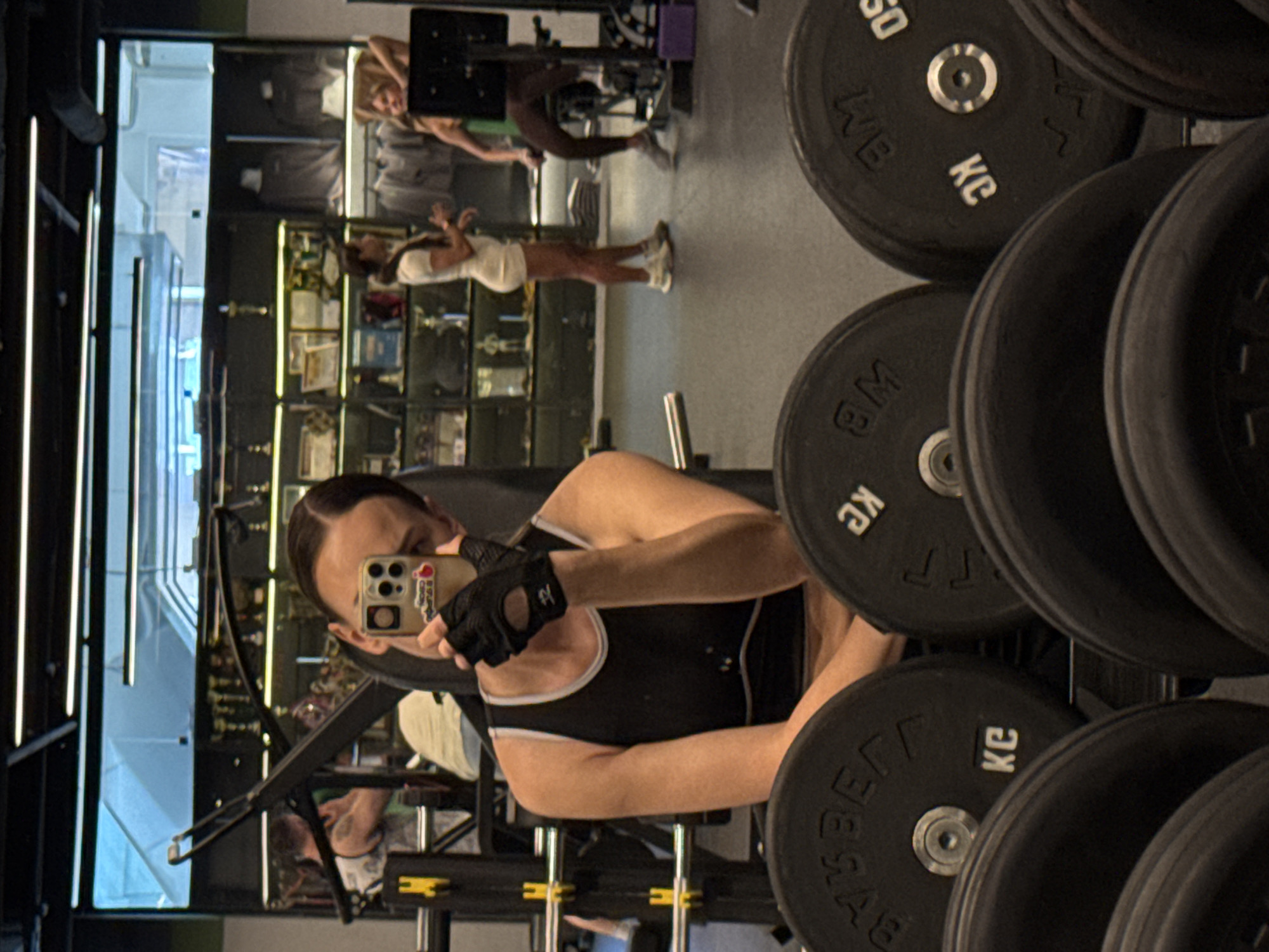 The image displays a mirror selfie taken in a well-equipped gym. The primary subject, a person with neatly parted dark hair, is seen from the chest up, wearing a black tank top with white trim and black fingerless gym gloves. They hold a smartphone with a light-colored case adorned with multiple colorful stickers or charms, partially obscuring their face as they capture the reflection.
To the right side of the frame, a stack of black barbell weight plates is prominently featured. Visible text on these plates includes "KG" (kilograms) and numerical values like "50" and "8," along with partial letters such as "M" and "B."
The background, reflected in the mirror, shows other gym patrons and interior details. To the left, bright vertical strip lights illuminate a section of the gym, possibly a doorway or passage. In the mid-ground of the reflection, another person in a light-colored top is partially visible behind the main subject. Further back, a woman dressed in a white athletic top and shorts is standing, and another person with blonde hair is seen bending over a piece of gym equipment. Along the far back wall, a large display unit holds numerous trophies, awards, or framed certificates, indicating a facility with a history of achievements.
The activity taking place is a self-portrait captured during a workout or break at the gym. The scene suggests an active, modern fitness environment with ample lighting. The visible text on the weights confirms the use of kilograms for measurement. The location context indicates this gym is in Sochi, Russia.