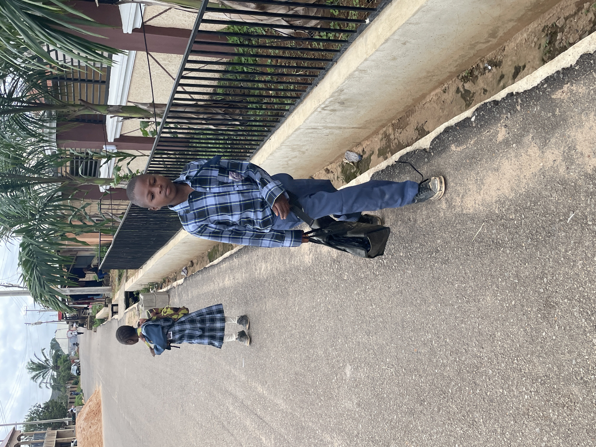 In Gura Top, Nigeria, two children in school uniforms walk along an asphalt road. The child in the foreground, a boy, wears a blue and white plaid shirt and dark blue trousers. He carries a black bag in his right hand. The second child, a girl, walks ahead of him, wearing a blue and white plaid dress over white leggings and carrying a backpack. They are walking on the right side of the road, with a concrete drainage ditch and a black metal fence to their right. In the background, palm trees line the street, and buildings are visible. The sky is overcast.