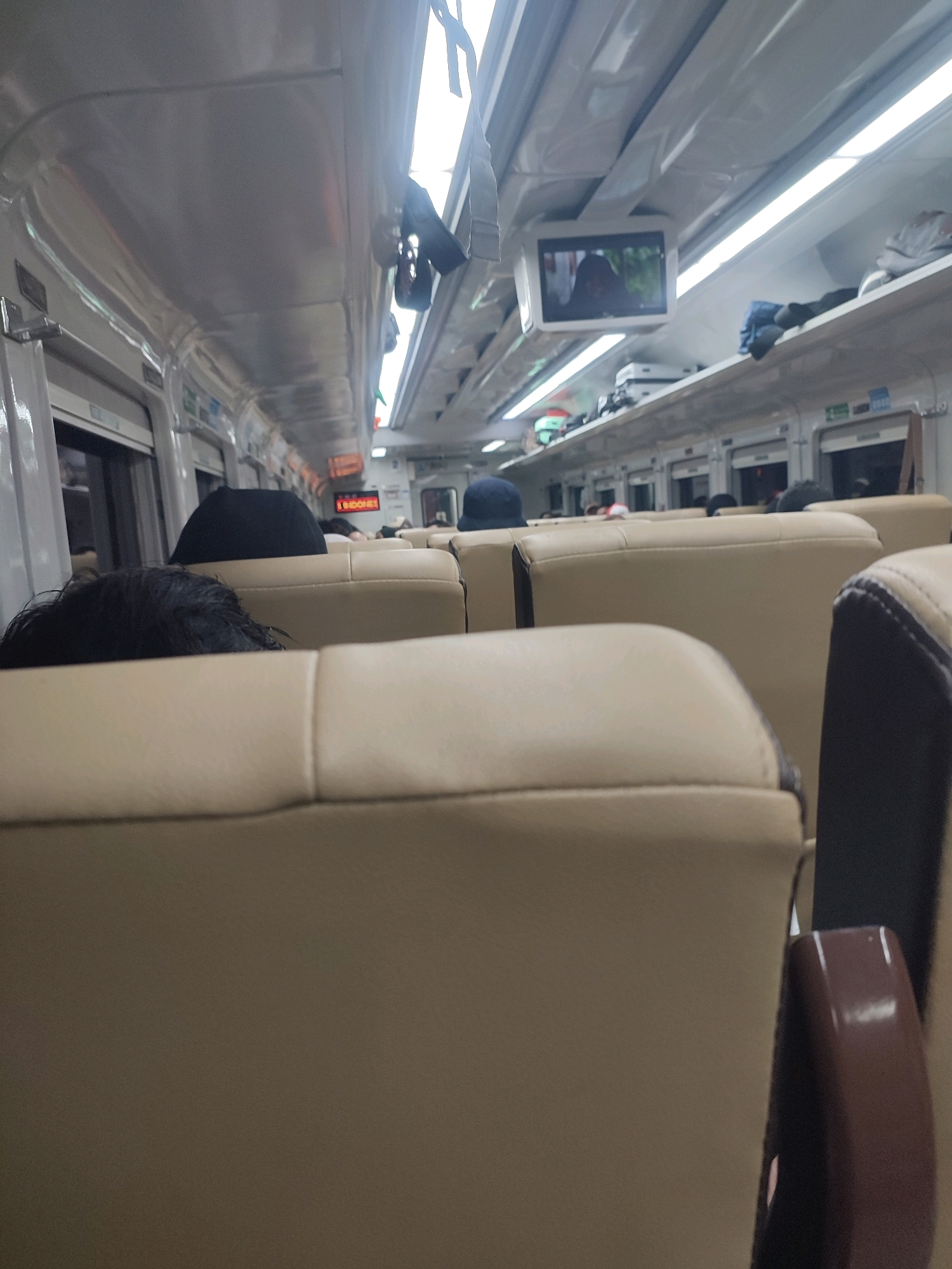 The image depicts the interior of a train carriage. The seating appears to be arranged in a side-by-side configuration with beige upholstery. The ceiling of the carriage is visible, featuring lighting fixtures and a small screen mounted above the seats, displaying a muted image. Luggage and personal items are stowed on overhead shelves. The perspective suggests the photograph was taken from a seat within the carriage, looking forward. People are visible, mostly their heads and backs, seated in the beige chairs. Their attire is not clearly discernible. The overall environment is typical of public transportation. There are no indicators to pinpoint the specific location within Arjawinangun, Indonesia, beyond it being inside a train.