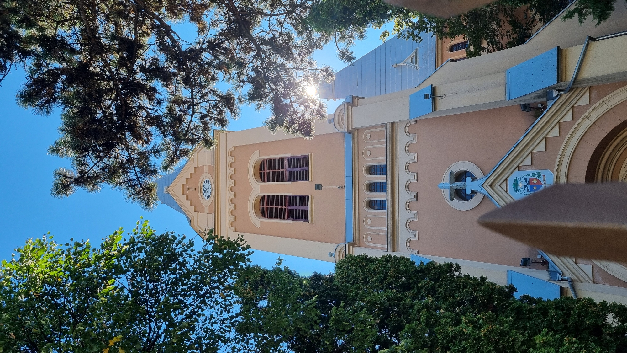 The image shows the bell tower of the Church of the Ascension of the Lord in Mukachevo, Ukraine, seen from a low angle through trees. The church has a peach-colored facade with cream-colored decorative elements. The bell tower is topped with a pointed spire. In the foreground, a blurred object, possibly a fence or a decorative element, partially obstructs the view of the church entrance. The sky is a clear blue. The sun is shining, with its rays visible through the branches of trees on either side of the tower.