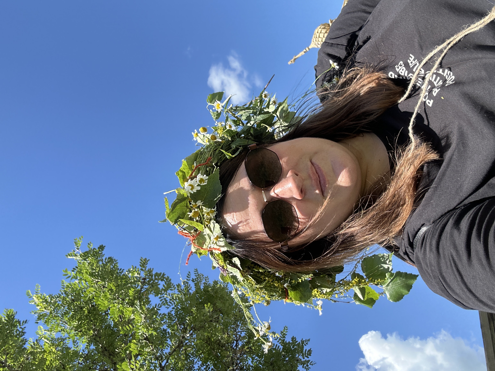 A low-angle, eye-level medium shot captures a woman from the chest up, wearing a large, wild flower and leaf crown. She is outside on a bright, sunny day, with a clear blue sky and scattered white clouds filling the background. To the upper left of the frame, the green leaves and branches of a tree are visible. The woman has medium-length brown hair and is wearing large, dark sunglasses. Her black t-shirt has white text on it that reads "POWERED BY POSITIVE LIFE". A coarse, natural-fiber string is draped across the front of her shirt. The overall atmosphere is casual and outdoor-oriented, suggesting a festive or celebratory occasion, possibly related to nature or a folk tradition. The location of Bolsherechye, Russia is not visually indicated in the image.