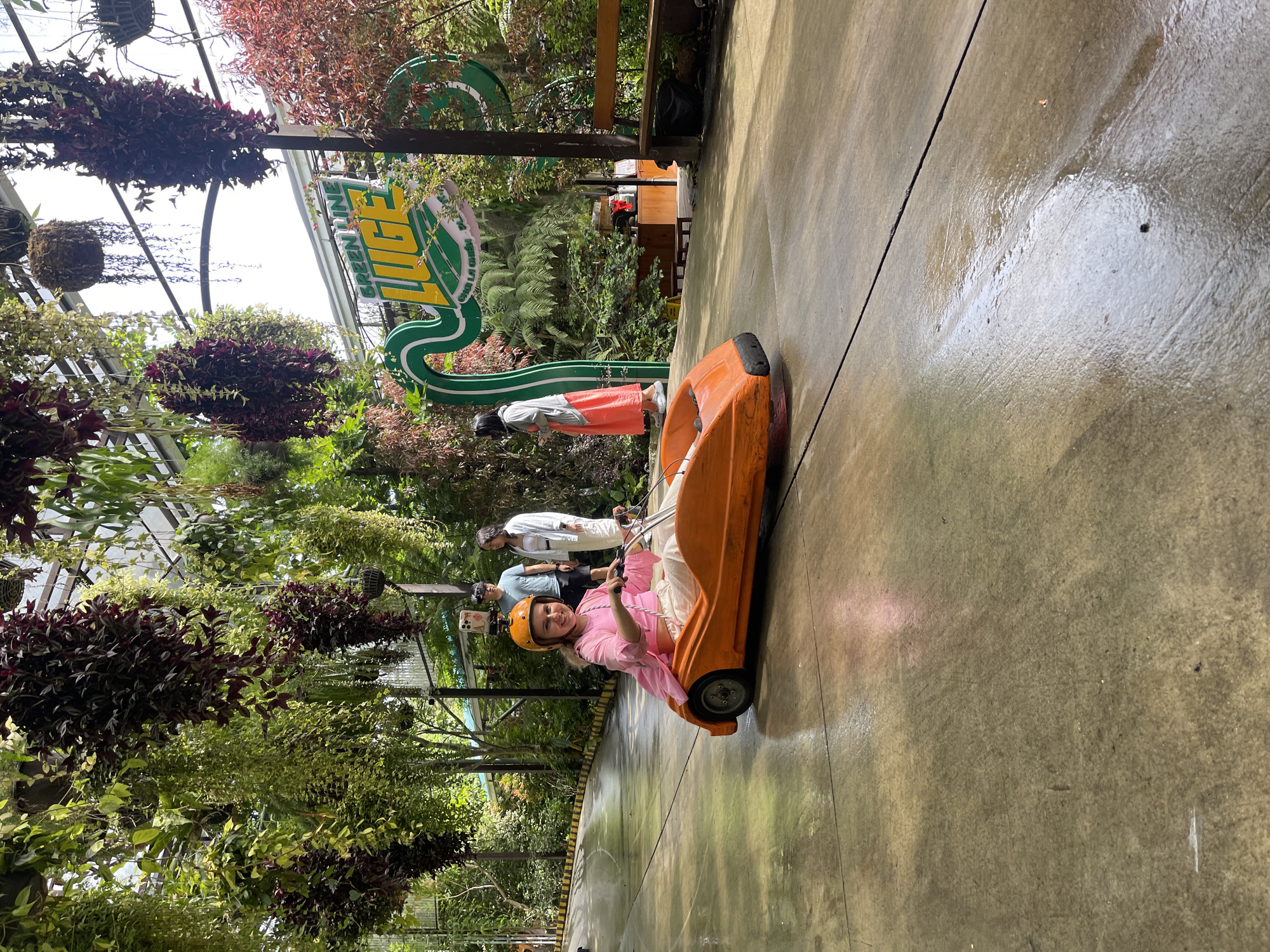 A young woman wearing a pink shirt and helmet is riding in an orange luge car at the Datanla New Alpine Coaster in Thôn 5, Vietnam. Other people are standing nearby, and the area is surrounded by lush greenery and hanging plants. A large sign reading "LUGE" is visible in the background. The ground appears to be wet.