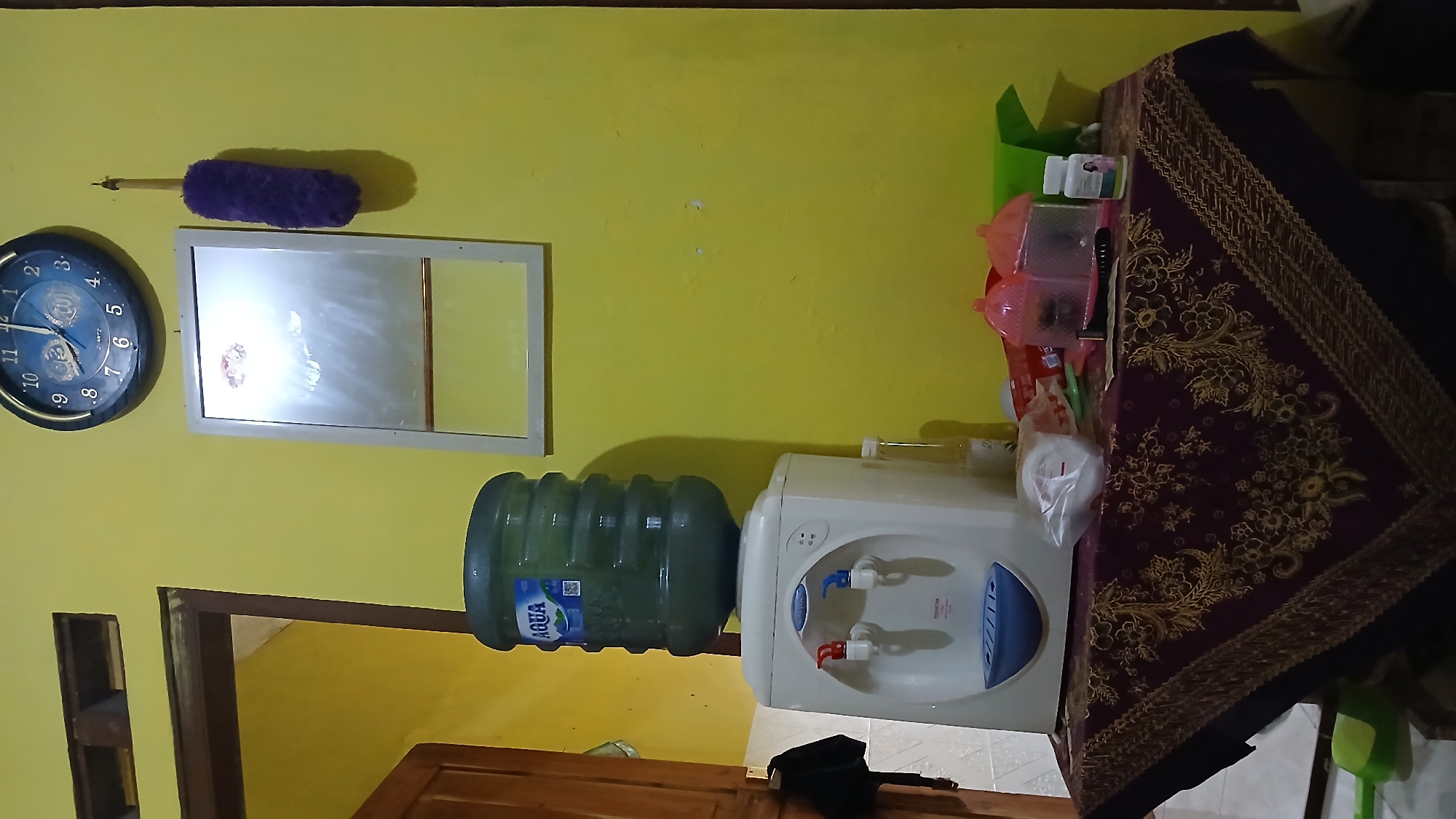 In the city of Mendut, Indonesia, a domestic scene unfolds within a room illuminated by artificial light against a vibrant yellow wall. Dominating the foreground is a white water cooler, its top adorned with a large, green Aqua brand water jug. To the right of the water cooler, a table is draped with a dark, patterned cloth, featuring intricate floral designs in gold and purple. On the table, various items are scattered, including a pink plastic container, a bottle of medication, a dark-handled grooming tool, and a plastic bag containing white disposable cups. To the left of the water cooler, a doorway leads to another area, with a wooden door partially ajar. Above the water cooler and table, a circular wall clock displays the time as approximately 11:15. To the right of the clock, a purple duster hangs from a hook on the wall. A rectangular mirror with a thin, light-colored frame is mounted on the wall between the clock and the water cooler. The floor is partially visible, revealing a green plastic item.