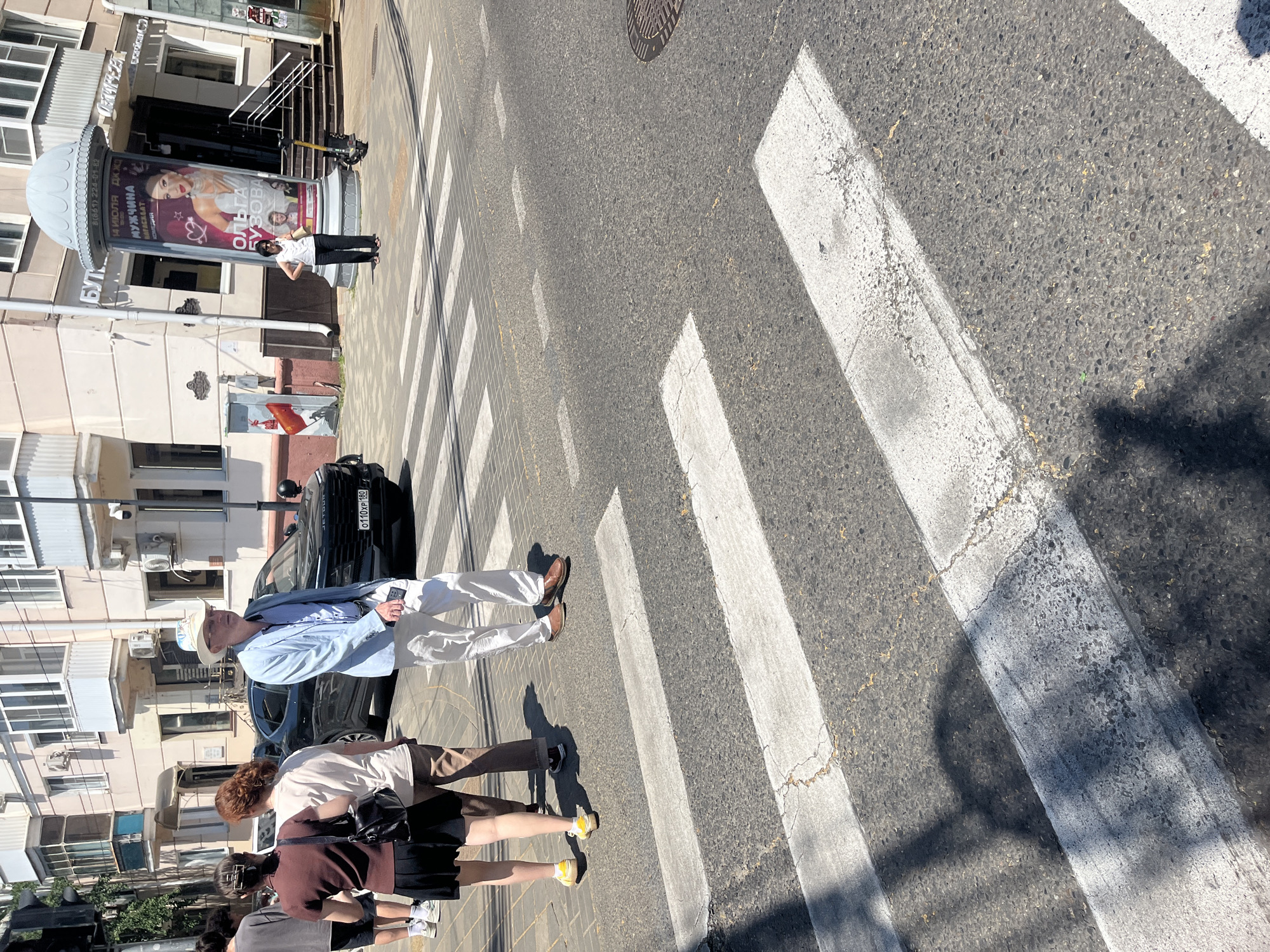 The image captures a street scene in Krasnodar, Russia, specifically on a sunny day at a pedestrian crossing. In the foreground, the white stripes of the crosswalk are clearly visible on the asphalt. A man in a light blue blazer, white pants, and a light-colored fedora is walking across the street, holding a phone in his right hand. Behind him, a group of three women are also crossing or waiting to cross. One woman in a dark top and black skirt, another in a beige t-shirt and brown pants, and a third person partially obscured. A black car with a license plate reading "0110XP 180" is stopped at the edge of the crosswalk. To the right of the street, a building with multiple windows and balconies stands, and a tall cylindrical advertisement pillar displays an image of a woman. A yellow scooter is parked near the pillar. The overall scene depicts an everyday urban environment with pedestrians and traffic.