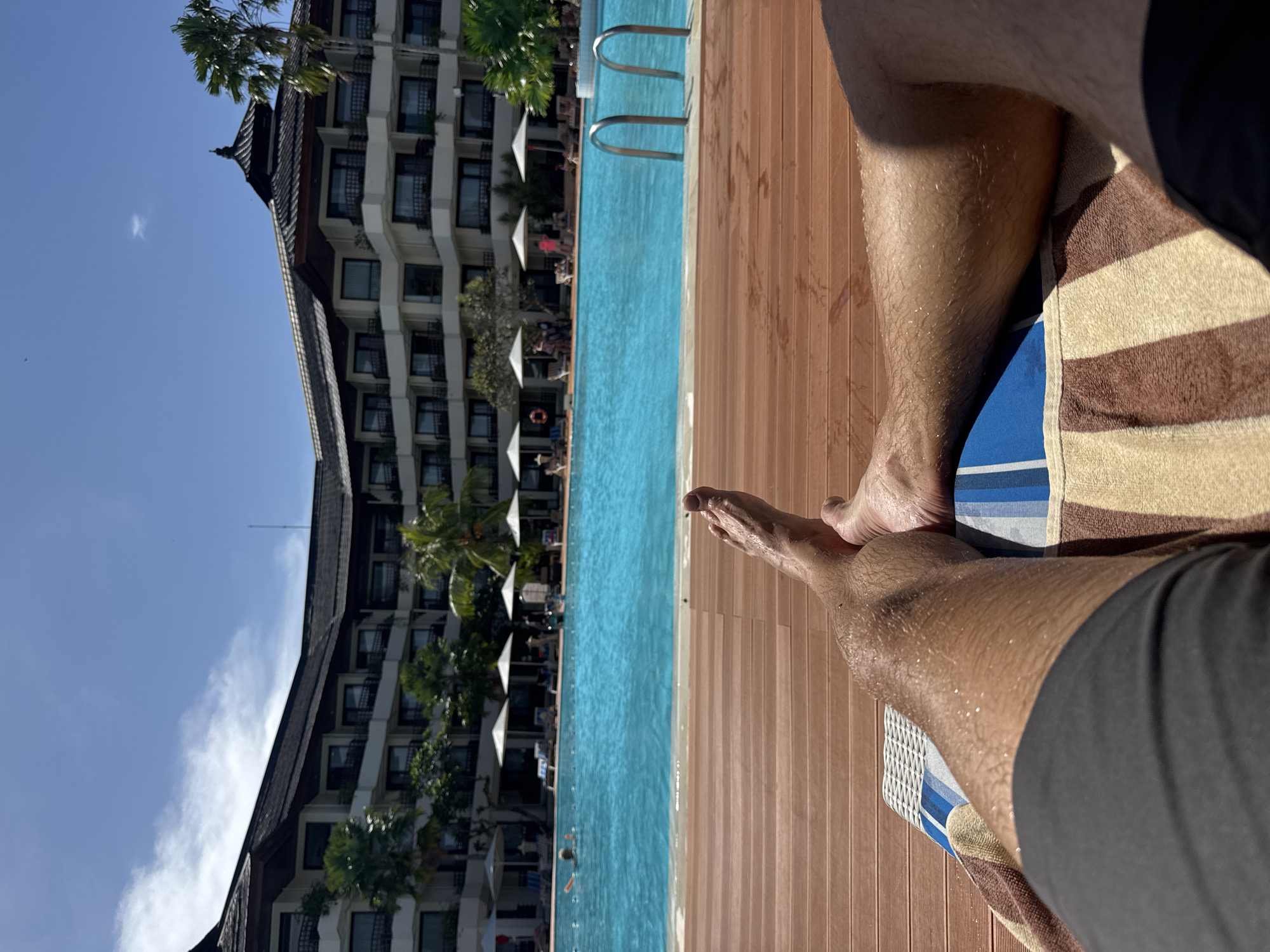 This media file captures a first-person perspective of a relaxing scene at a resort swimming pool, set in Denpasar, Indonesia. The image, viewed upright, shows a person's wet, tanned legs and feet resting on a brown and beige striped towel, which is placed over a blue and white striped lounge cushion. The foreground features wooden decking that runs alongside a clear, azure swimming pool.
In the midground, the inviting blue pool contains several individuals swimming, with two metal ladders providing access. White sun umbrellas are visible lining the far side of the pool, shading various lounge chairs where other guests are relaxing. Lush green palm trees are scattered around the pool area, adding to the tropical ambiance.
The background is dominated by a grand, multi-story hotel building with a distinctive traditional tiered, dark roof, possibly reflecting local Balinese architecture. The building features numerous balconies. Above the hotel and palm trees, the sky is a bright, clear blue with a few wispy white clouds, indicating sunny weather. The scene is bathed in strong sunlight, suggesting it's daytime, likely mid-morning or afternoon. The overall impression is one of leisure, vacation, and tropical relaxation. There is no visible text in the image.