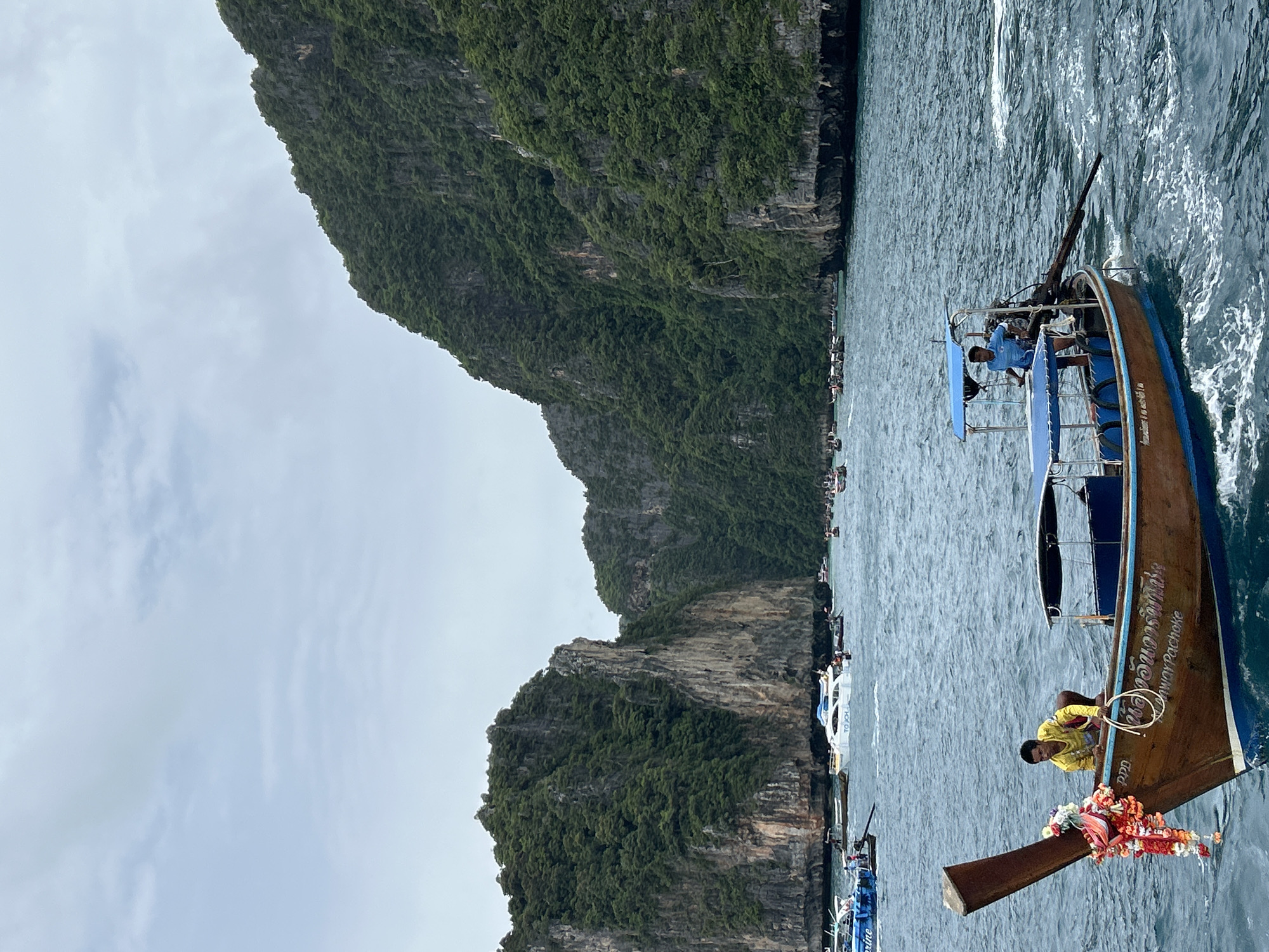 This image depicts a scene in Ao Nang, Thailand, featuring a longtail boat in the foreground. The boat is wooden with blue trim and has a canopy. Two men are visible on the boat; one, wearing a yellow shirt, is leaning over the front of the boat, and another, in a blue shirt, is standing near the engine at the back. The boat is in motion, creating ripples on the water. In the background, several other boats, including a white yacht, are scattered across the sea. Towering limestone cliffs covered in lush green vegetation dominate the background, characteristic of the karst landscape of Ao Nang. The sky is overcast with a few clouds.