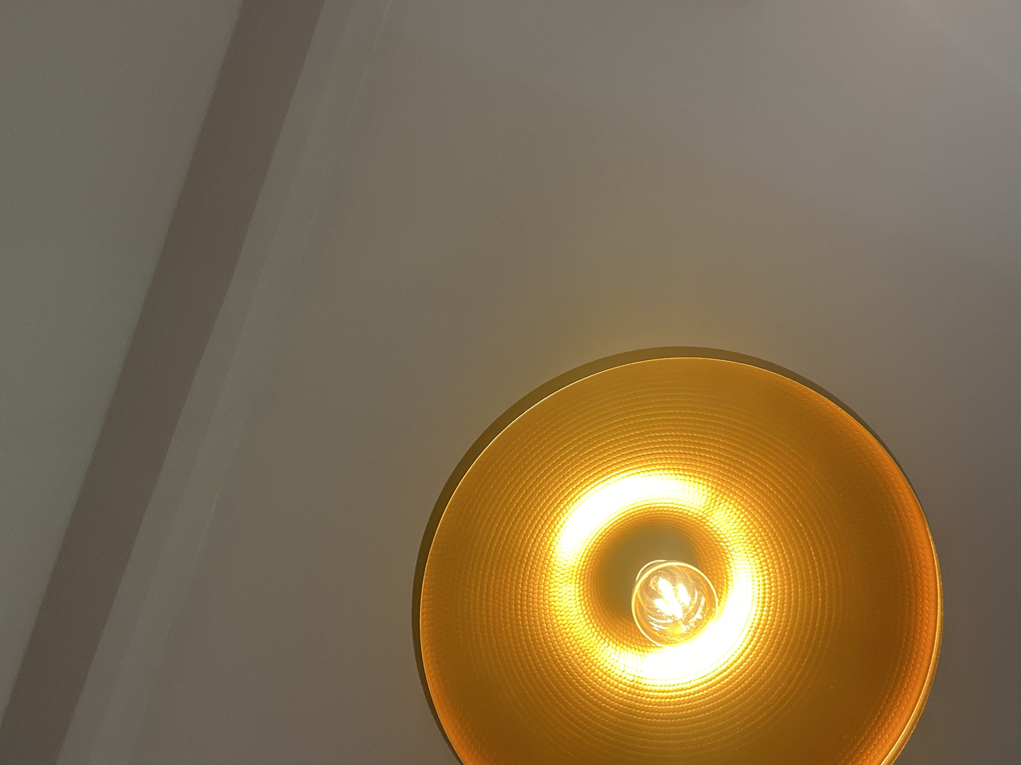 A close-up, downward-facing shot captures the interior of a golden pendant light fixture, emitting a bright, warm glow. The lightbulb, a classic filament design, is clearly visible at the center of the circular reflector. The textured interior surface of the lamp, likely made of metal, displays a repeating pattern of small circles. The background consists of a neutral-colored ceiling, with a subtle shadow line diagonally across it, suggesting an architectural feature or the angle of overhead lighting. There are no people or identifiable objects other than the light fixture itself and the ceiling. The image provides no discernible details about its specific location within Budva, Montenegro.