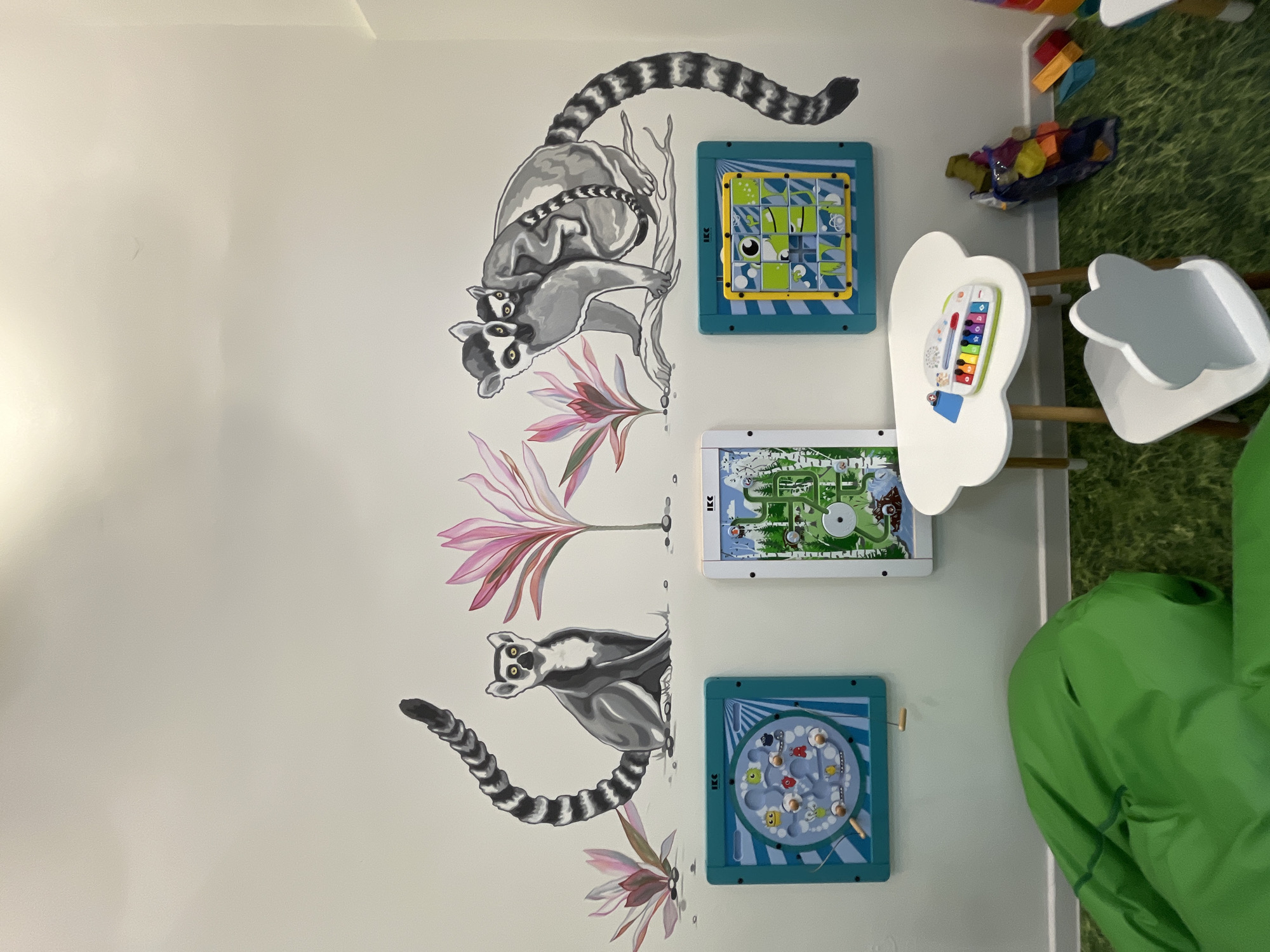 The image shows a wall in what appears to be a playroom, possibly in a childcare facility or home setting. On the wall, there are two lemur murals with pink stylized flowers, interspersed with three mounted interactive game boards. One board contains a sliding puzzle, another features a marble maze, and the third shows a small, indoor playground. To the right of the wall is a small, cloud-shaped table and chair set, and there is a green beanbag chair. The floor is covered in a grass-patterned mat. Without further context, it is impossible to verify this location is in Chaikovsky, Russia.