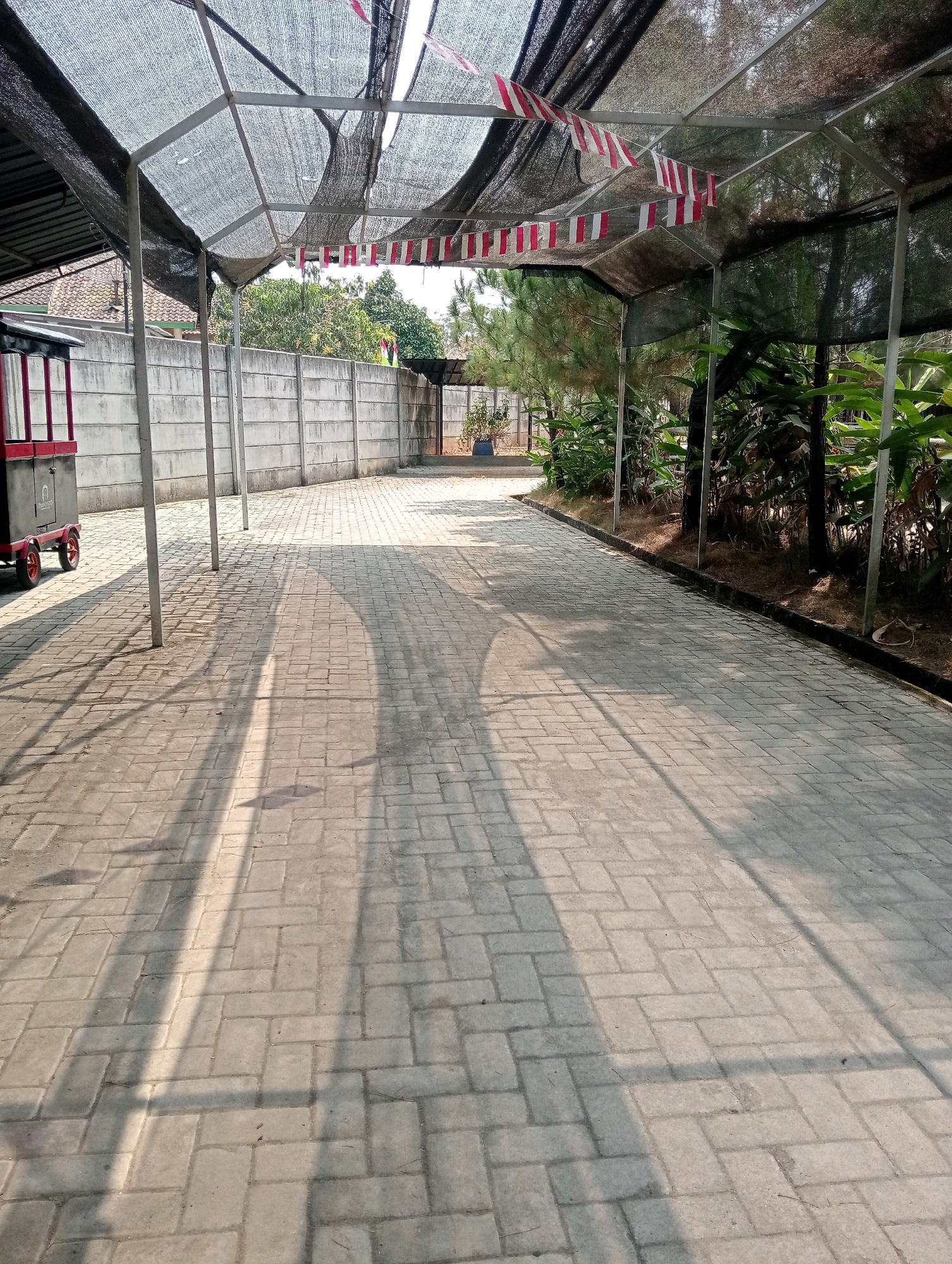 An outdoor paved walkway runs under a shaded canopy, extending into the distance. The walkway is constructed of interlocking rectangular pavers laid in a herringbone pattern, showing signs of wear and tear. The canopy, made of a black mesh material, is supported by metal poles and creates dappled shadows on the ground. Red and white triangular flags are strung across the canopy in a festive arrangement. To the left, a section of a red and black cart with wheels is visible, partially obscured by a metal pole. Beyond the walkway, a concrete wall stretches across the scene, forming a boundary. Trees and lush green foliage are visible behind the wall and under the canopy, suggesting a natural setting. The sunlight casts long shadows from the canopy's supports and foliage onto the paved surface. The environment appears to be a public space or an outdoor venue. The image was taken in Pringsewu, Indonesia.