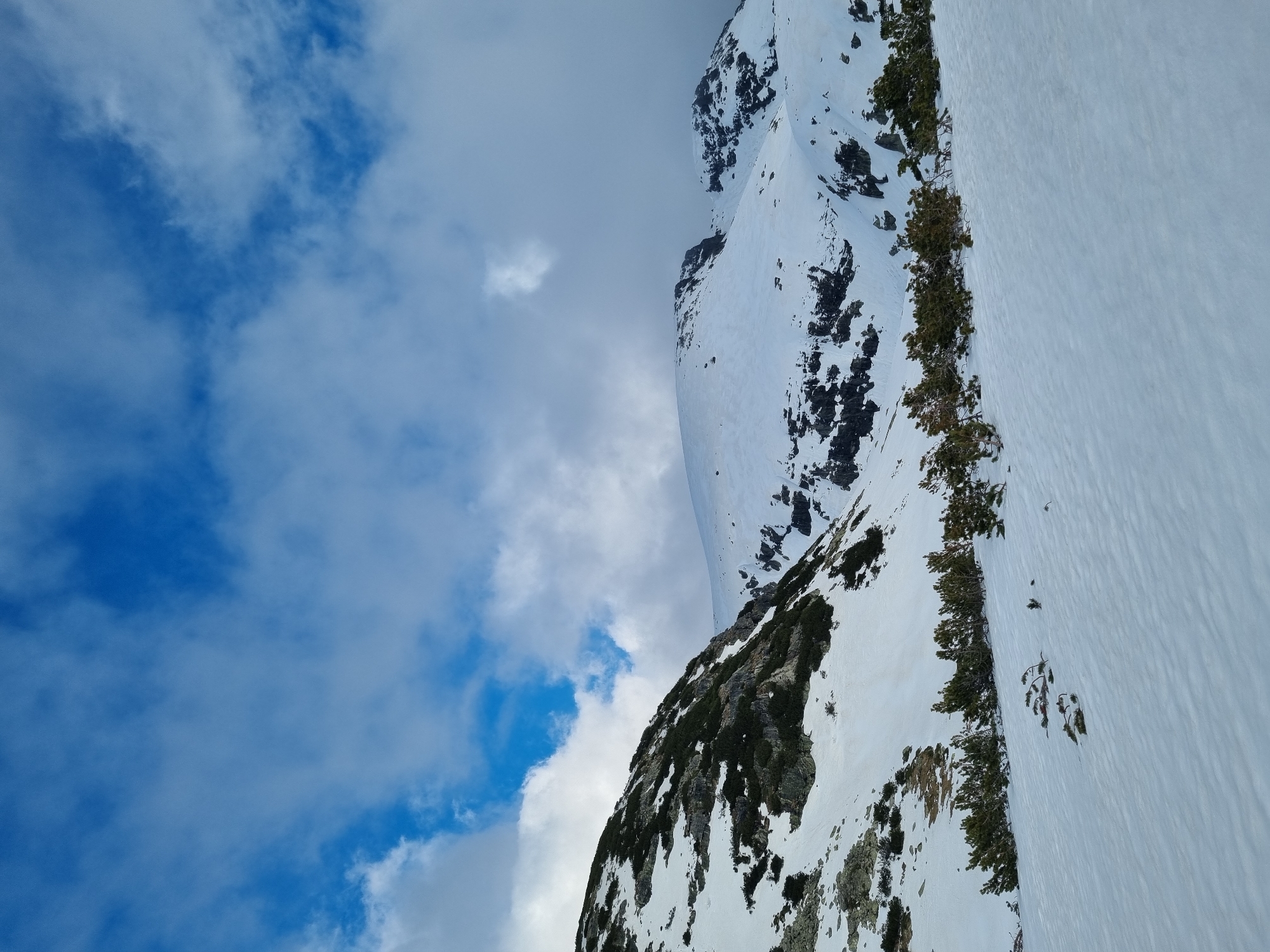 The image captures a mountain landscape in Govedartsi, Bulgaria, partially covered in snow. The sky is visible with patches of blue and white clouds. A rocky, snow-covered mountain slope ascends into the sky. Patches of green vegetation punctuate the snow cover. The composition of the image is such that the mountain appears to be viewed from a low angle.