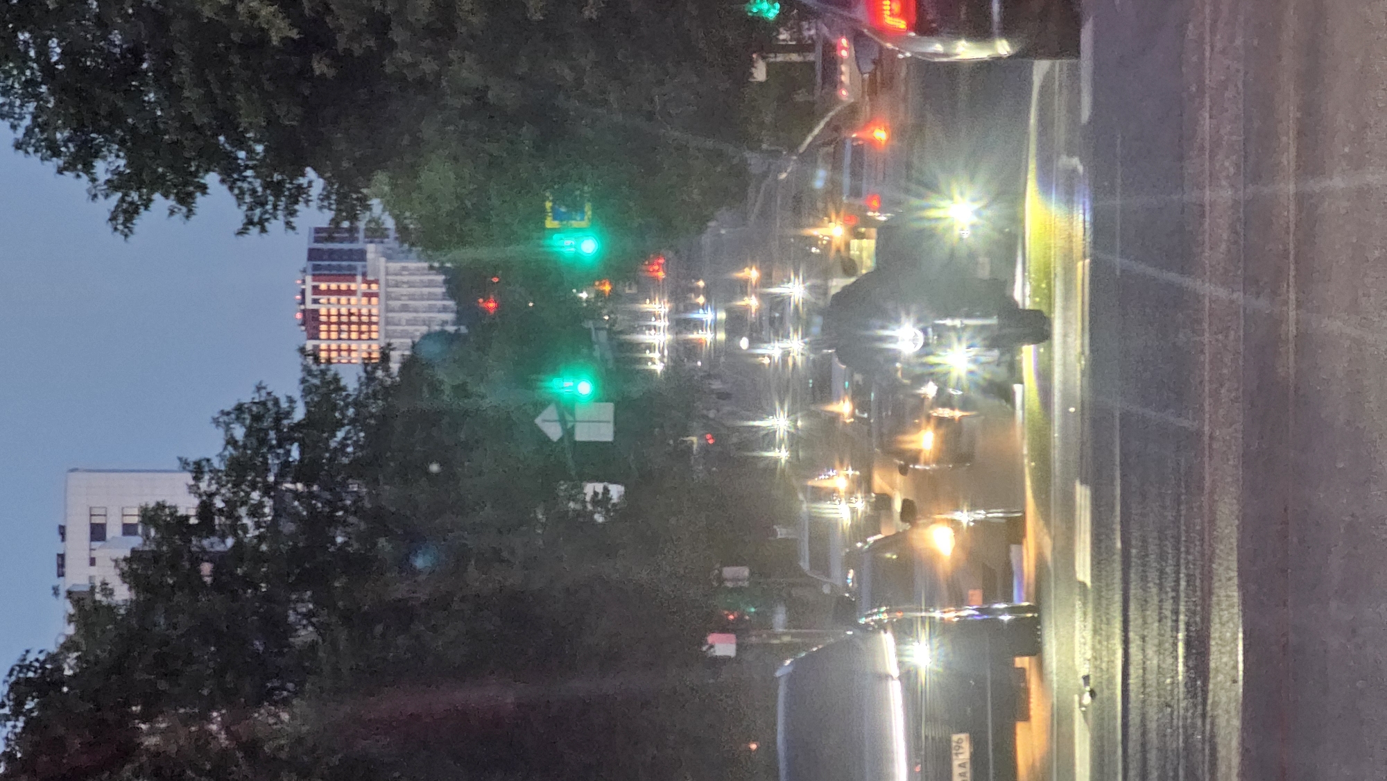 The image captures a street scene at dusk or dawn in Yekaterinburg, Russia, likely at an intersection. Tall trees line both sides of the road, their dark foliage contrasting with the dim sky and the illuminated cityscape in the background. In the distance, modern high-rise buildings are visible, one of them illuminated with a grid of orange lights, suggesting it is an apartment building or hotel.
The foreground is dominated by traffic. Several cars are stopped or moving slowly, their headlights and taillights creating streaks of light on the wet asphalt, indicating recent rain. A prominent motorcycle, its headlights shining brightly, is positioned in the center of the road, also with its lights creating lens flares. The traffic lights are green, allowing for movement. A person is visible on the motorcycle, silhouetted by the bright headlights. The overall atmosphere is one of urban transit in low light conditions, with the wet road reflecting the artificial lights of the vehicles and traffic signals. The specific location appears to be a main thoroughfare with multiple lanes of traffic.