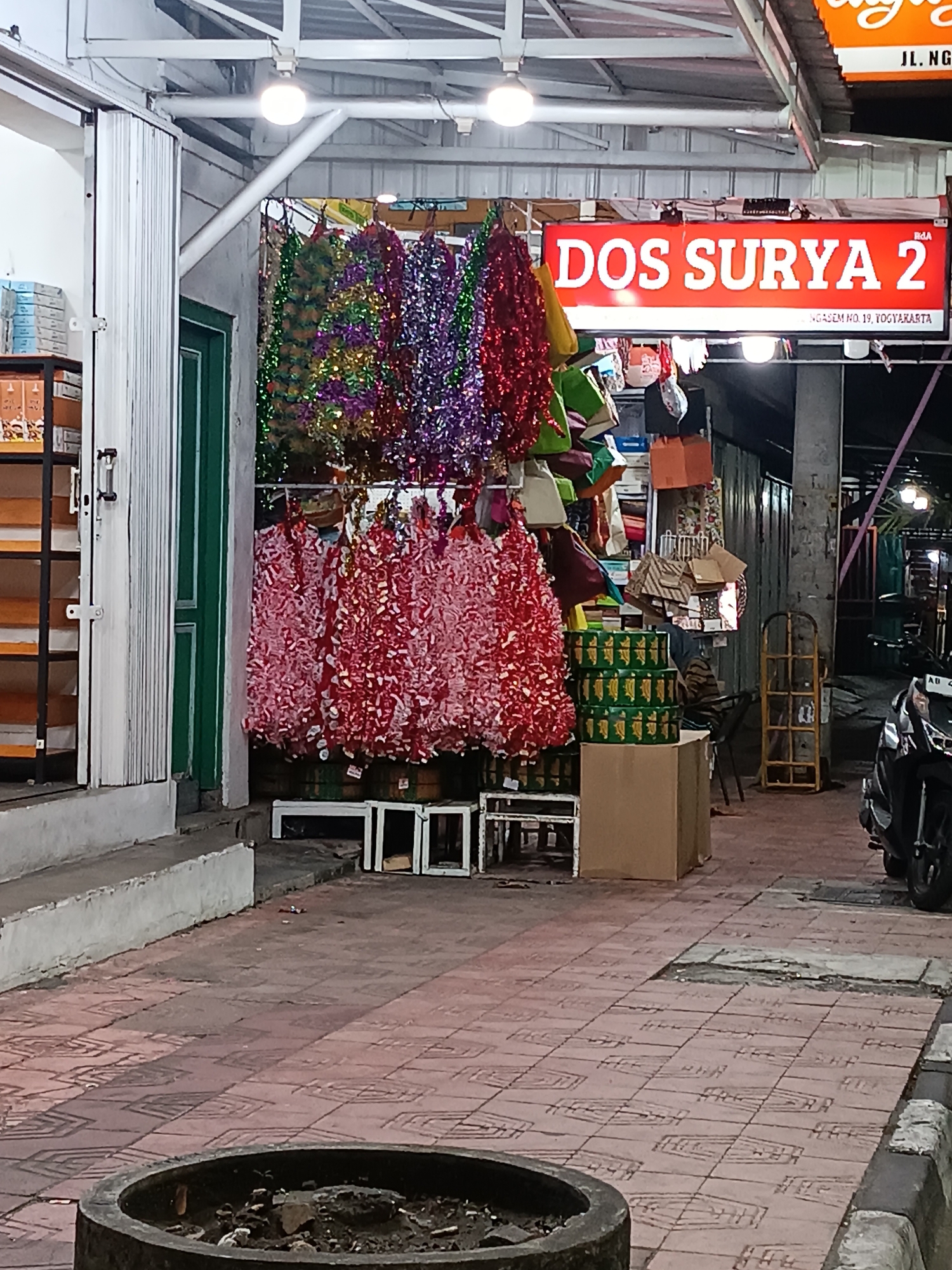 This is a street-level view of a shop in Yogyakarta, Indonesia. The shop appears to sell party decorations, with colorful tinsel hanging outside. A prominent red sign reads "DOS SURYA 2" in white letters. Below this sign, smaller text indicates "NGASEM NO. 19, YOGYAKARTA". The storefront features a white roll-up door and a green door. In front of the shop, there are stacks of green boxes and some cardboard boxes. A motorcycle is parked on the right side of the image. The ground is paved with reddish-brown tiles. The scene is lit by overhead lights, suggesting it is nighttime or dusk. There are no people visibly present in the scene.