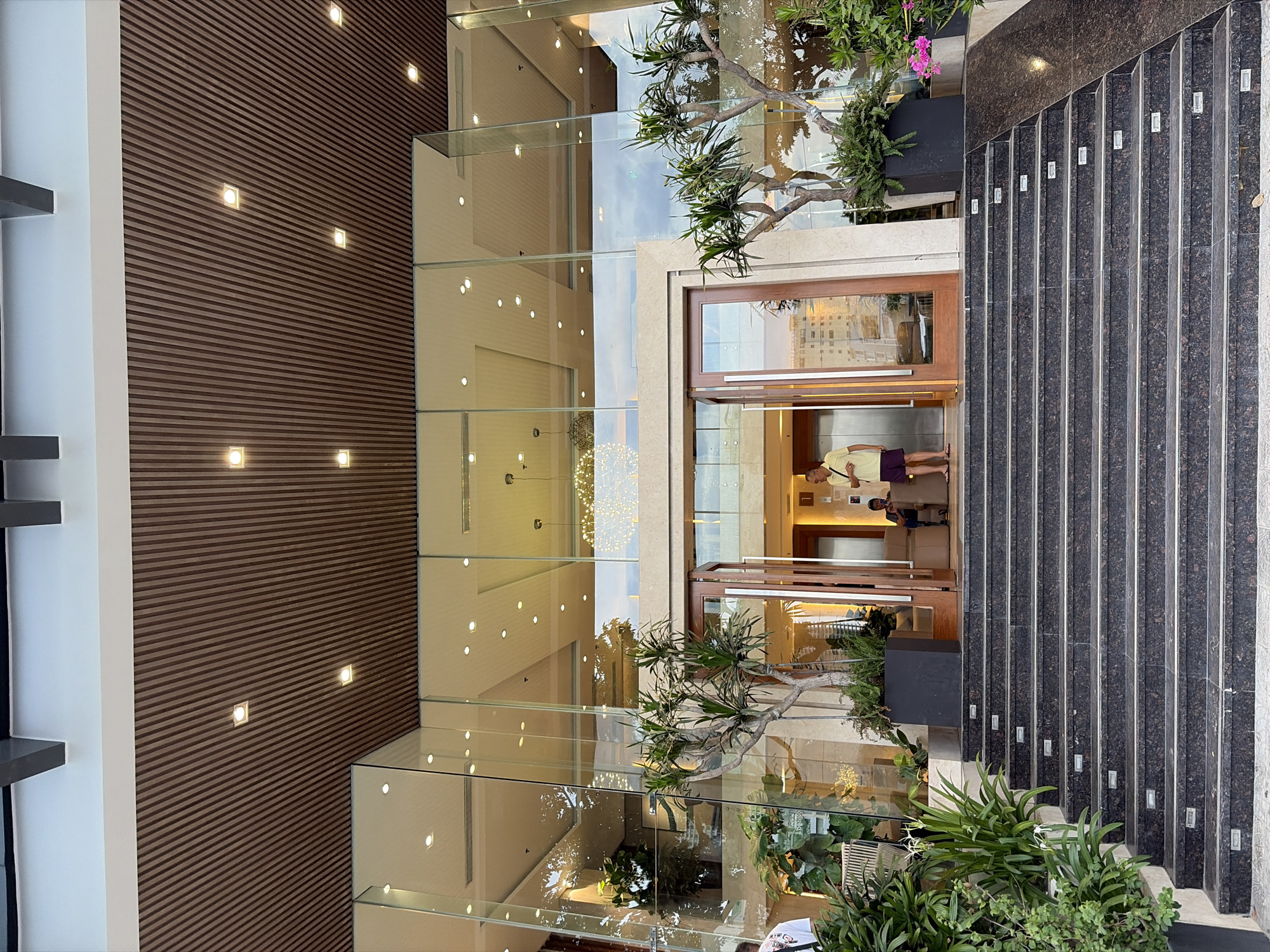The image shows the entrance to a modern building with large glass windows and a wooden slatted ceiling. A grand staircase made of dark speckled stone leads up to the entrance, which features double wooden doors and an elevator. Two potted plants, one with lush green foliage and the other with spiky leaves, flank the entrance. A person in a yellow shirt and purple shorts stands near the door, looking towards the camera. Another person is seated inside the building. The scene appears to be in Đà Nẵng, Vietnam, based on the architecture and outdoor setting.