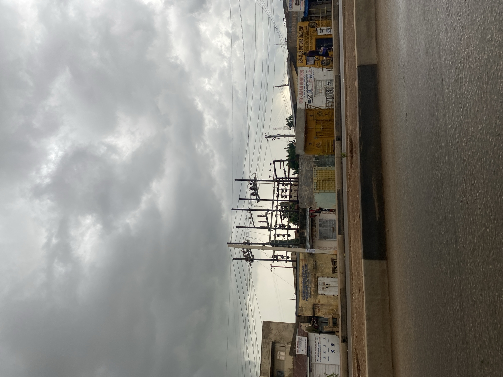 The image shows a street scene in Gura Top, Nigeria, under a heavily overcast sky. In the foreground, a wet asphalt road is visible, with a concrete curb separating it from a dirt embankment. Beyond the embankment, a row of shops and buildings line the street. Overhead, a complex network of electrical wires and utility poles dominate the sky. The buildings appear to be a mix of commercial establishments. One building has signage indicating "CHY-JAPAN VENTURES" and "BENJASS MOTORS (NIG) ENT." Another building to the left has signage that is partially obscured but appears to be an advertisement with multiple logos. The buildings have varied facades, some weathered and discolored, with closed shutters and doors. A few individuals are visible in front of the shops, and one person is walking along the sidewalk. The overall impression is of a busy urban environment, likely a market or commercial district, during a cloudy day.