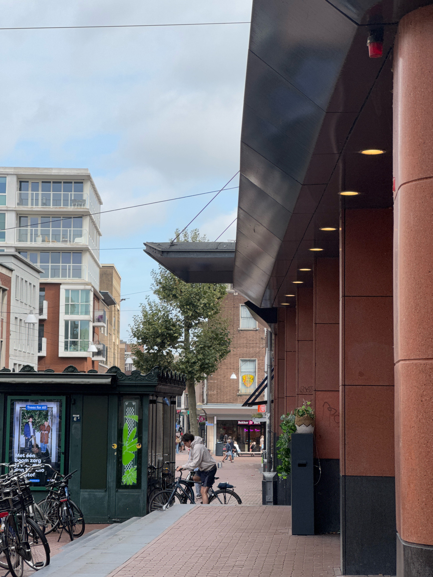 The image captures a street scene in Nijmegen, Netherlands, on a cloudy day. In the foreground, a person wearing a light-colored hoodie and dark shorts is standing next to a bicycle, appearing to adjust it. They are positioned on a paved walkway in front of a dark green, kiosk-like structure with posters displayed. Several bicycles are parked to the left of the kiosk.
To the right, a building with a modern facade featuring large reddish-brown stone-like panels and a dark sloping overhang dominates the view. The overhang has a series of recessed lights. A red fire alarm is visible near the top of the building's facade.
In the background, various buildings are visible, including a contemporary multi-story structure with balconies and large windows. Further down the street, commercial establishments are present, one of which is labeled "Bakker Bart." Trees and utility wires are also part of the urban landscape. The ground is paved with reddish-brown bricks.