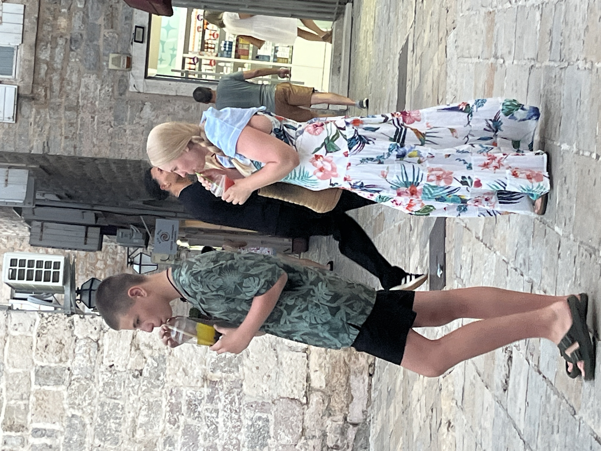 A young person with short hair, wearing a green patterned short-sleeved shirt and black shorts, is drinking from a clear plastic cup with a straw. They are standing on a cobblestone street in Budva, Montenegro, next to a stone building. To their right, a woman with blonde hair braided into pigtails, wearing a white floral maxi dress and a light blue head covering, is also drinking from a clear plastic cup with a straw. Behind them, other people are walking in the street, and shops are visible in the background. The street appears to be a pedestrian area with shops lining one side.