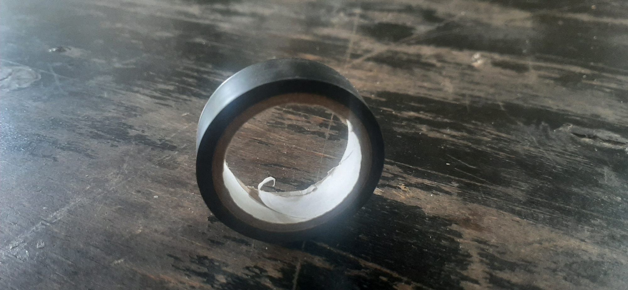 A roll of black electrical tape lies on a dark, weathered wooden surface. The tape is partially unrolled, revealing a white backing. The surface appears to be a table or countertop with visible wood grain and some imperfections, suggesting an indoor setting. The lighting is even, casting no strong shadows, which might indicate an overcast day or artificial lighting. There are no people or animals visible in the image, and no discernible text. The scene is simple and focused on the object itself. The overall impression is one of everyday utility and a slightly rustic aesthetic due to the wooden surface.