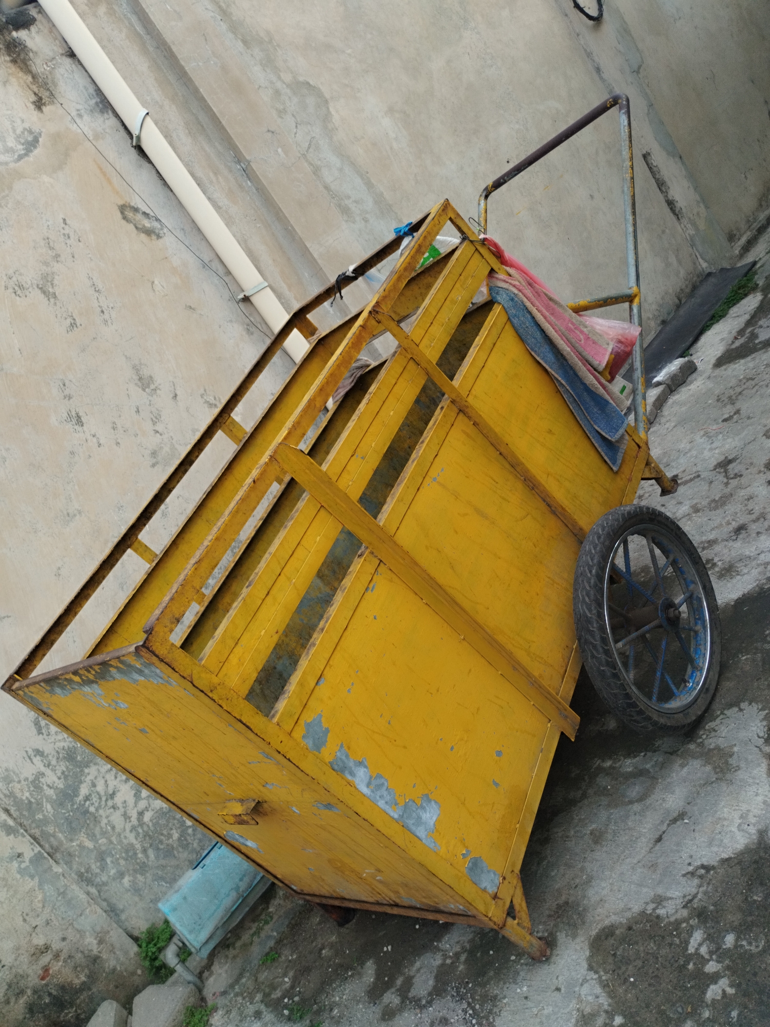 The image depicts a yellow, metal cart with a single wheel, positioned on a concrete surface. The cart has a weathered appearance with peeling paint. A metal pipe runs vertically along a textured, concrete wall in the background. There are some blue and pink fabrics draped over the top of the cart. The overall scene appears to be in an outdoor urban setting, likely in Surabaya, Indonesia, given the context. There are no people or animals visible in the frame, and no discernible actions are taking place. The lighting suggests it is daytime.