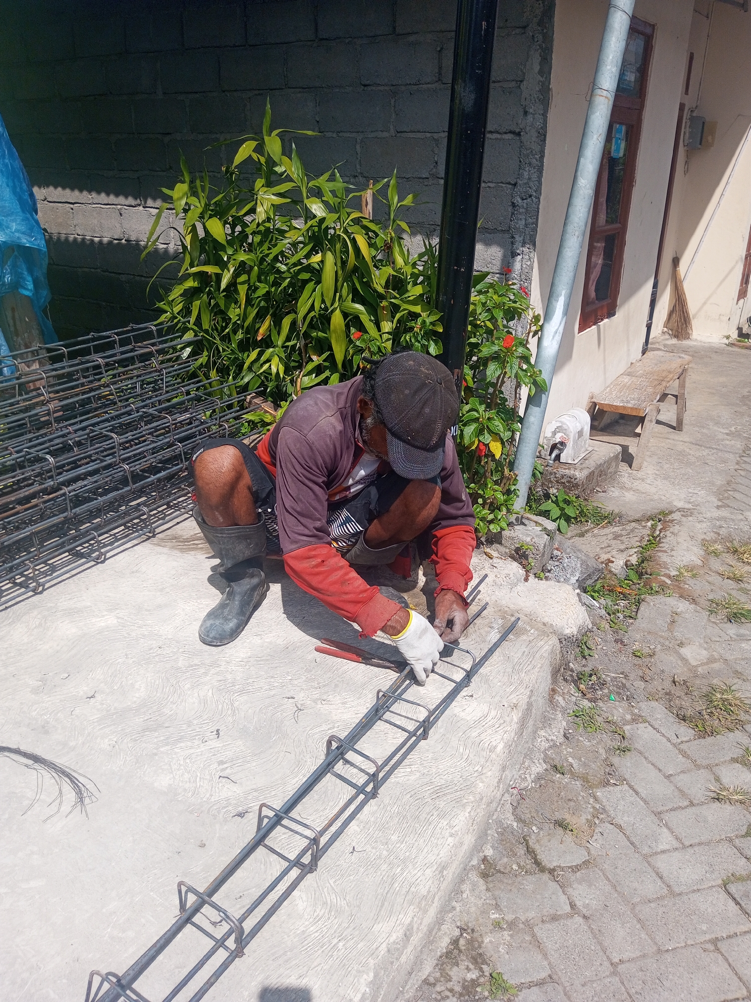In Andongsili, Indonesia, a construction worker kneels on a concrete surface, assembling rebar. He wears a dark baseball cap, a maroon shirt with red sleeves, black shorts, and black rubber boots. His hands are protected by white gloves, and he is actively working on a metal structure composed of long bars and reinforcing loops. To his left, a large stack of pre-assembled rebar sections is visible. Behind him, a grey brick wall casts a shadow, and lush green plants grow, some with small red flowers. A black vertical pole stands near the plants, and a silver pipe runs down the side of a building. The ground is a mix of concrete and brick pavers.