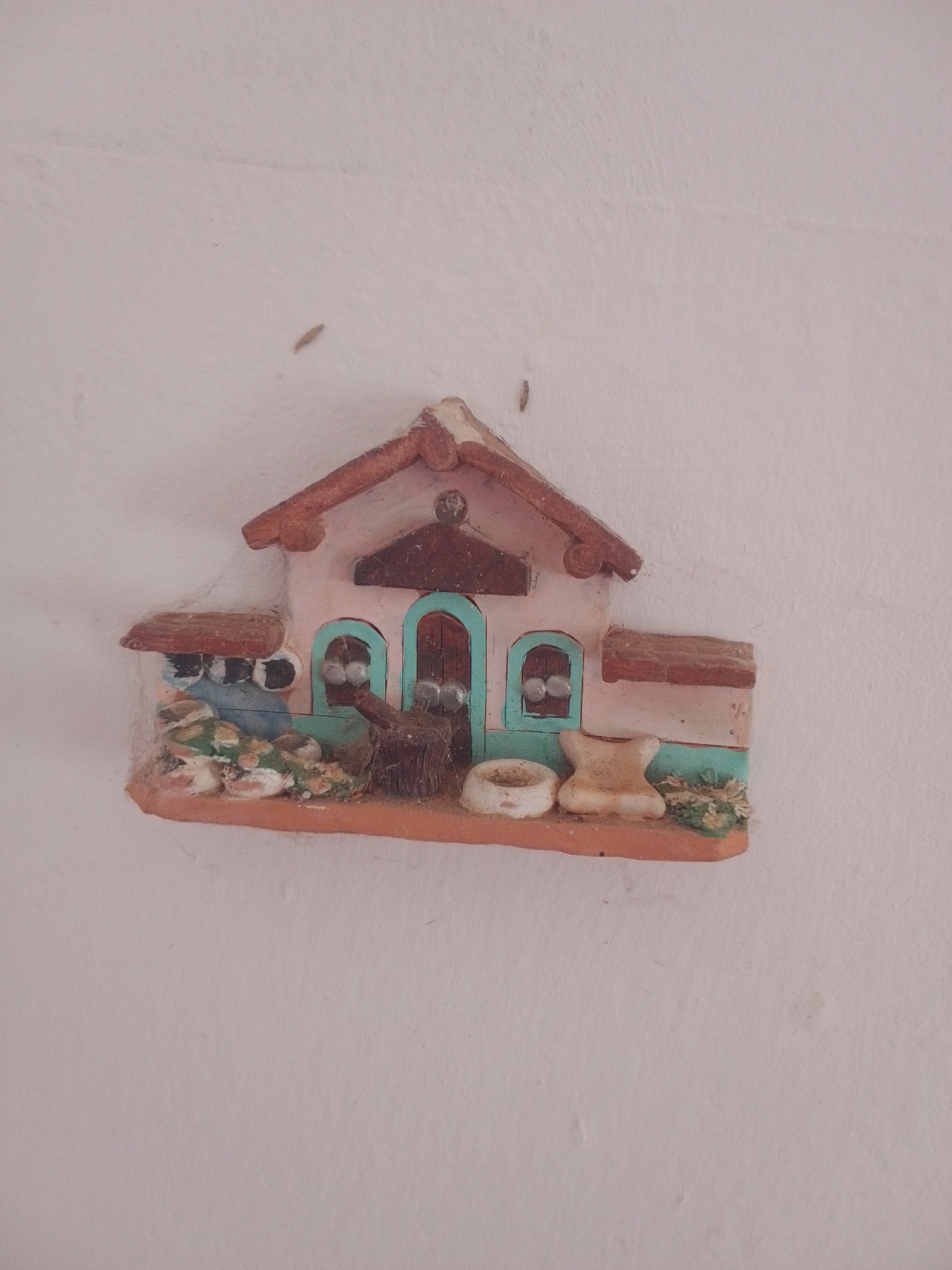 This media file depicts a small, decorative ceramic house ornament mounted on a textured, off-white wall. The house has a reddish-brown tiled roof and a light pink facade with teal-colored arched doorways and windows. In front of the house, there are miniature details such as a broom leaning against a wooden stump, a small white bowl, and a tan-colored object resembling a small stool or cushion. Green and brown elements suggest miniature landscaping or foliage. The scene is illuminated by soft, diffused light, suggesting an indoor setting or an overcast day. Several small, dark, elongated objects, possibly insects or dust, are visible on the wall surrounding the ornament, and a thin layer of cobweb is attached to the left side of the ornament. There is no visible text in the image. The context provided indicates the location as La Tagua, Venezuela. The overall impression is of a quaint, rustic decorative piece.