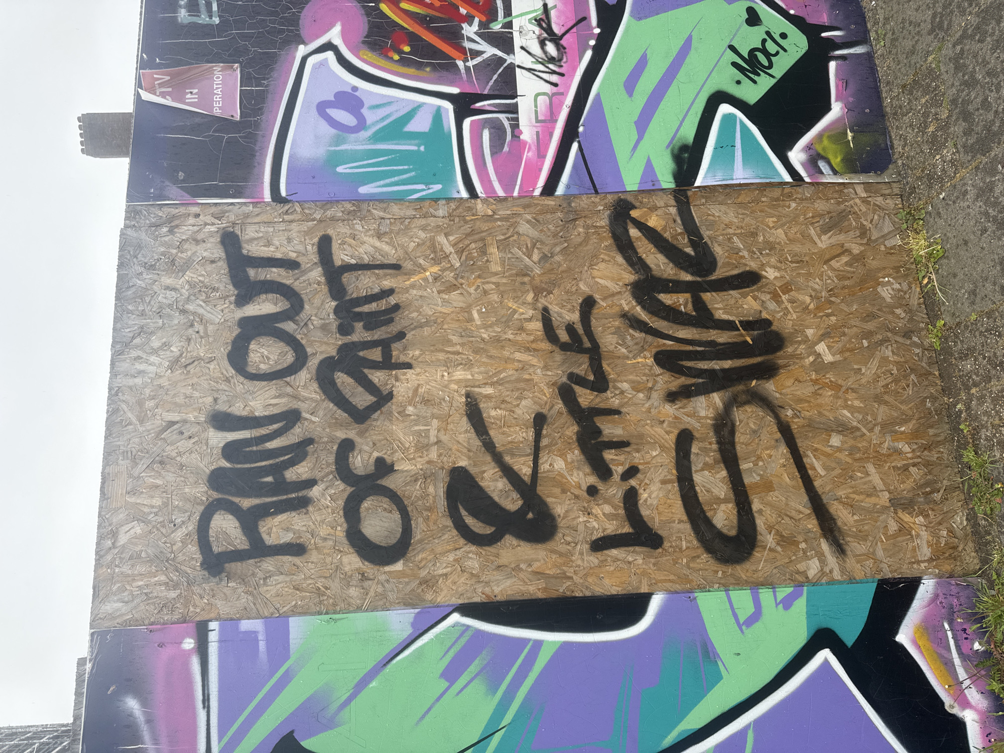 The image shows a section of wall or barrier in an urban environment, featuring layers of graffiti. At the top and bottom of the image are panels covered in colorful, abstract graffiti. A rectangular plywood board has been placed over a section of the existing graffiti. Written on the plywood in black spray paint are the phrases "RAN OUT OF ART" stacked vertically, and to the right "LITTLE SLUT" written in a dripping, cursive style. The background is slightly out of focus, suggesting an overcast day. Patches of grass are at the bottom right. The location is possibly in London, United Kingdom.