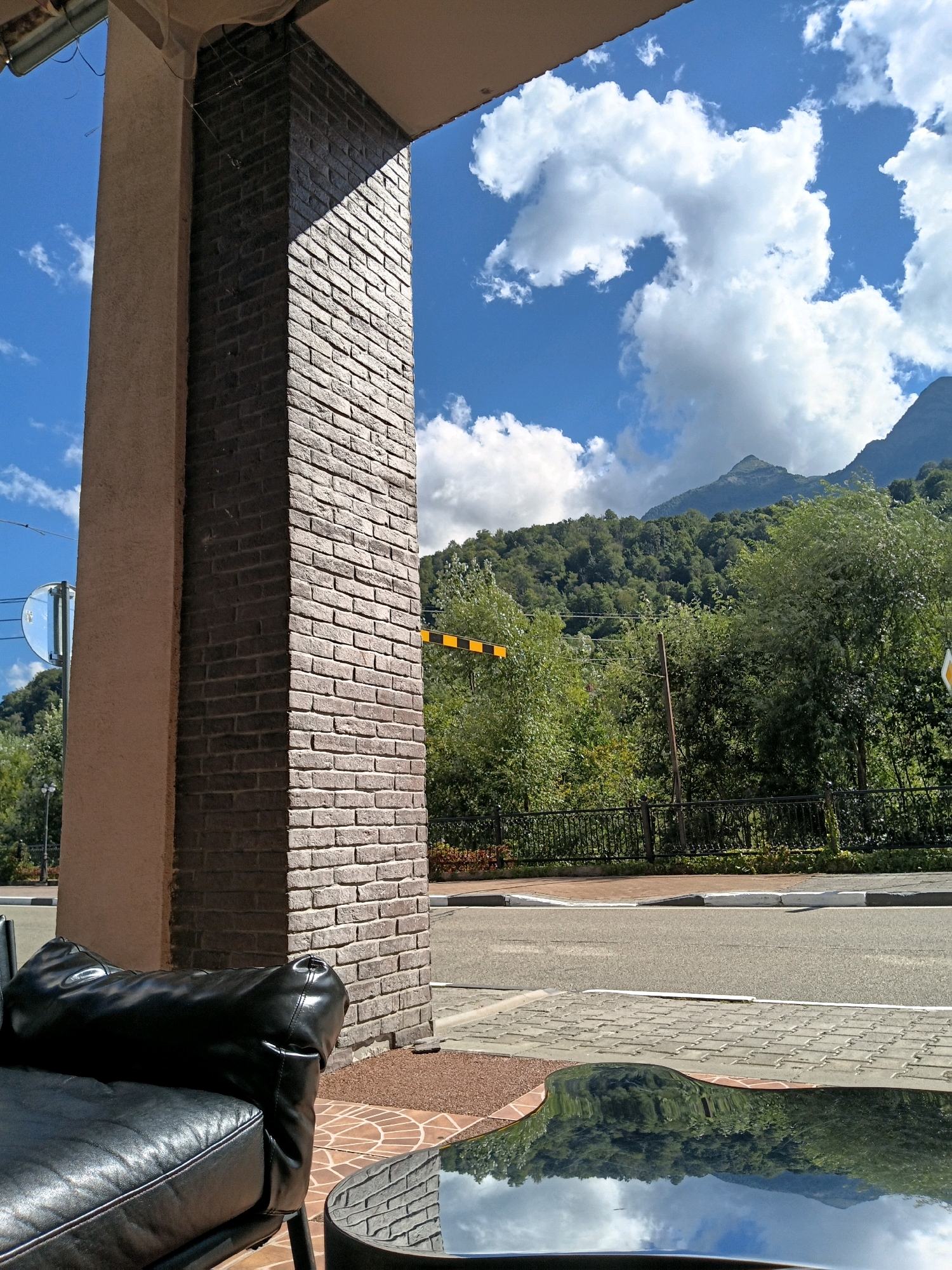 The image shows a view from a covered outdoor area, looking out towards a road and a mountainous, forested landscape under a partly cloudy sky. In the foreground, a black leather sofa is partially visible on the left, and a circular glass-topped table is in the lower right, reflecting the scene. A brick-clad pillar stands to the right of the sofa, casting a shadow. Beyond the covered area, a road runs horizontally, with a black metal fence and dense green trees lining the far side of the road. Mountains covered in trees rise in the background, with a peak visible against the sky. A yellow and black striped barrier is also visible near the road. The location is identified as Estosadok, Russia.