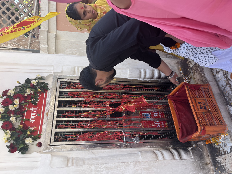 The image shows a man in black clothing bending down and dropping petals into an orange crate in front of a shrine. The shrine is adorned with red flowers and red and yellow ribbons. The shrine has a metal gate, which is covered in red strings and a padlock. Behind the man, there is a woman with a yellow headscarf, and a small child in a pink dress, also dropping petals into the crate. The man's face is obscured by his dark hair.  The shrine is located at 30.723 latitude and 76.86 longitude in Panchkula, India. The shrine is a place of worship and the people in the image are performing a ritual by dropping petals into the crate. The environment is dusty and there are some petals scattered around the crate. The orange crate is labeled "R+K BALAJI".