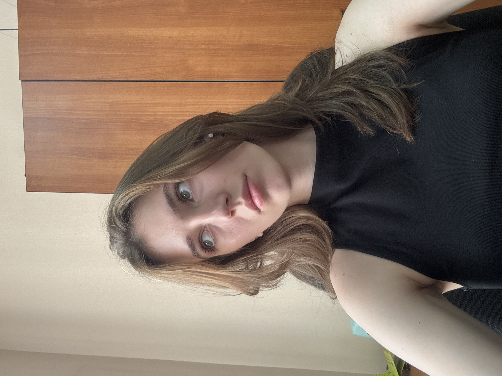 A young woman with light brown hair is pictured from the chest up, looking off to the left. She is wearing a black sleeveless top. Behind her, to the right, is a wooden wardrobe with two doors. To the left, a beige wall is visible. The image was taken in the city of Perm, Russia.