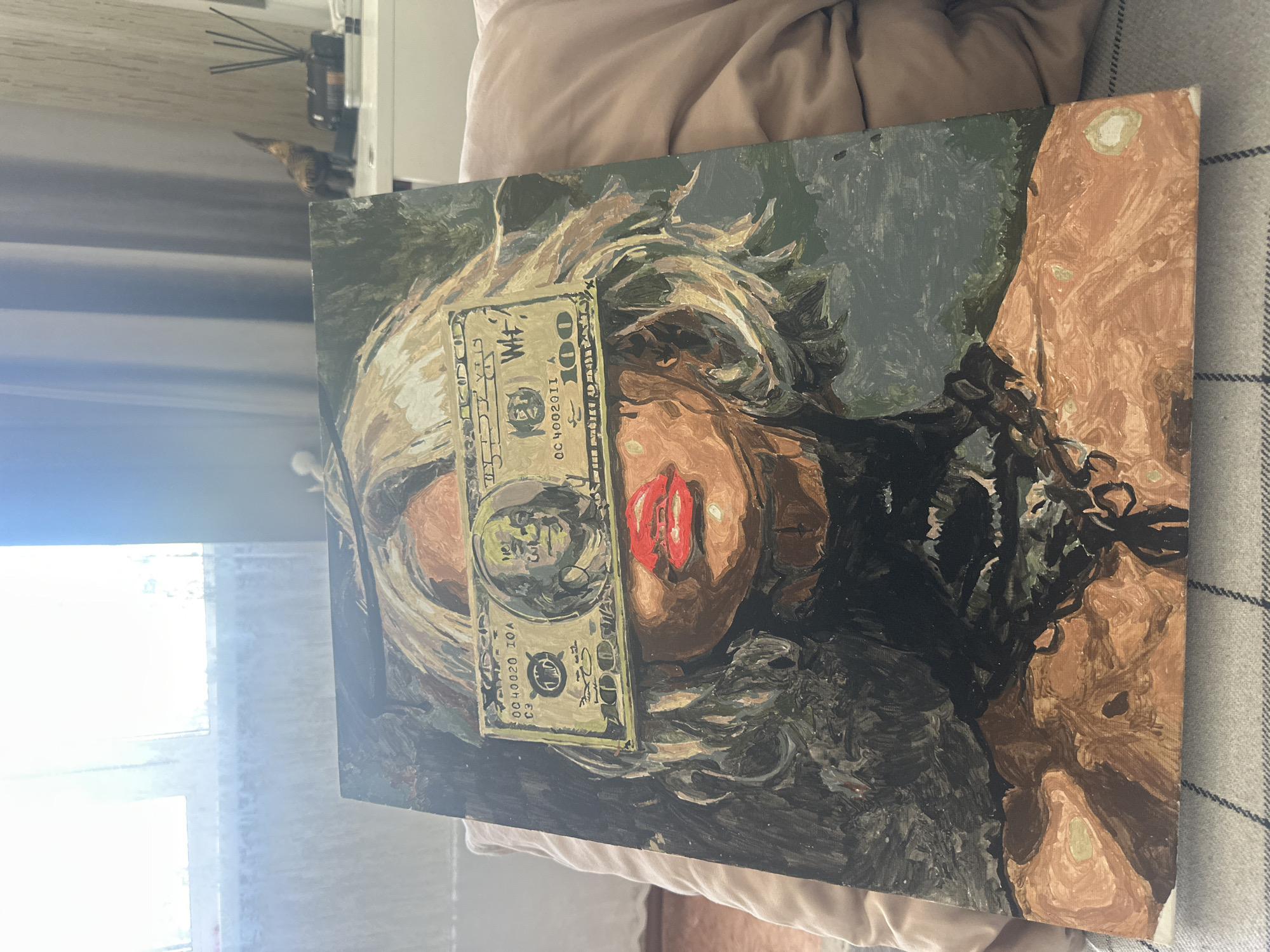 The image shows a painting resting on a plaid surface with a beige pillow in the background. The painting depicts a woman with a hundred-dollar bill obscuring her eyes. Her lips are painted a bright red, and the rest of her face is done in earth tones. Her hair is a mix of blonde and brown. The background of the painting is a dark green. Behind the painting are a window on the left, and a shelf with objects on the top left. The image does not contain any information to suggest the location is in the city of Высокий, Russia.