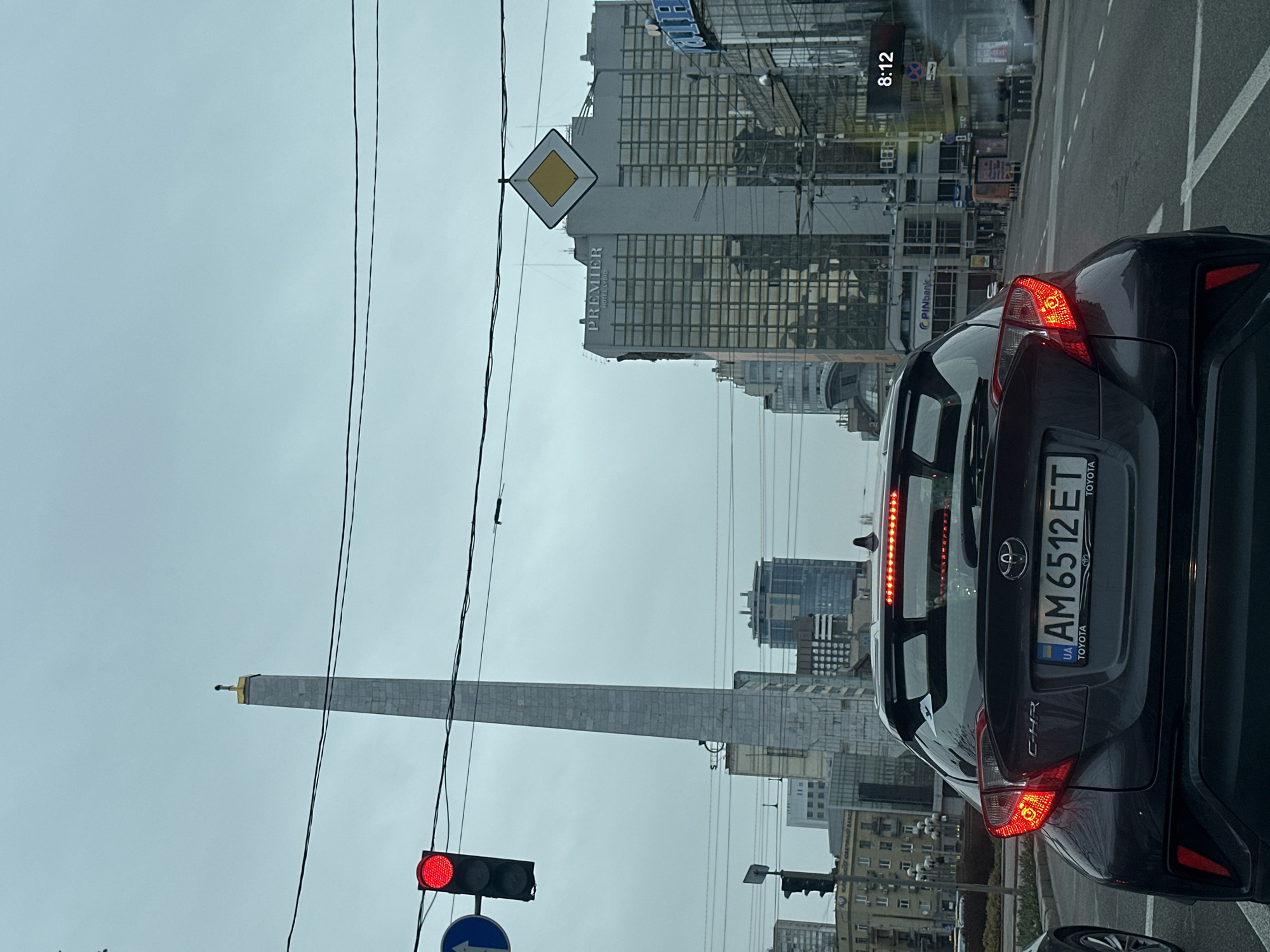 The image shows a gray Toyota C-HR with Ukrainian license plate AM 6512 ET driving in Kyiv, Ukraine. The Victory Monument is visible in the background. A diamond-shaped road sign and the Premier Hotel Lybid are also visible. The time on a nearby digital clock is 8:12. The sky is overcast.