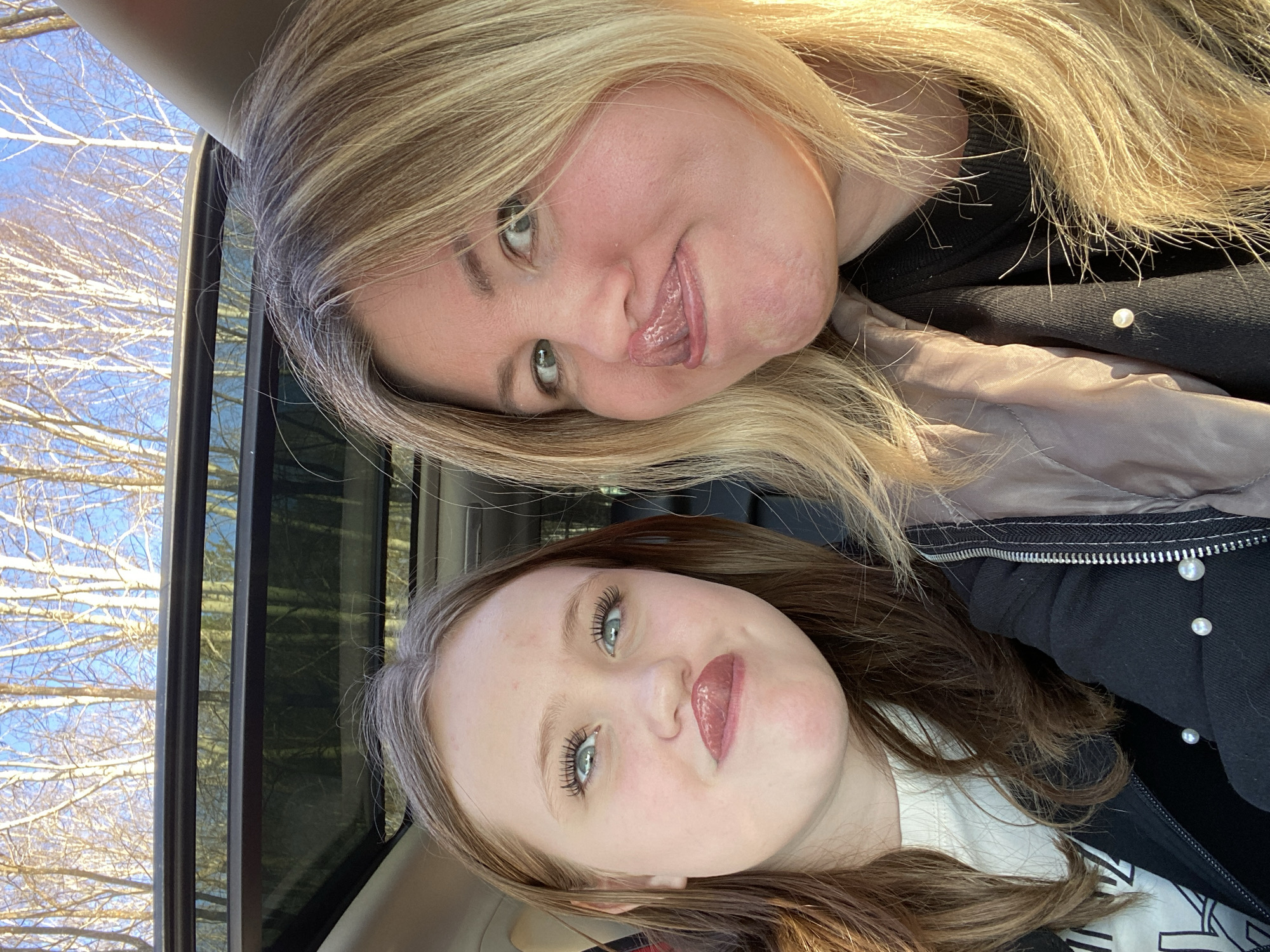 An indoor selfie captures two females, likely mother and daughter, inside a vehicle. The image is oriented sideways, with their heads tilted.
The upper subject is an adult woman with long, wavy blonde hair. She has fair skin, green eyes, and is making a playful expression with her tongue slightly extended. She wears a black top under a light brown or taupe jacket, with a visible pearl detail on her black top.
The lower subject is a younger female with long, wavy brown hair and fair skin. She has light blue-green eyes enhanced with mascara and wears pink lip gloss, offering a slight smile towards the camera. She is dressed in a white top beneath a black jacket featuring a zipper and multiple small, pearl-like buttons.
The setting appears to be the interior of a vehicle, with dark-tinted windows visible behind the subjects. Through the window, a bright blue sky is evident, along with numerous bare, light-colored tree branches, characteristic of a deciduous forest in late autumn or winter. This aligns with the given context of Chaikovsky, Russia, which experiences cold seasons.
The activity is a shared moment of taking a selfie, conveyed by their direct gaze at the camera. The expressions of both individuals suggest happiness, playfulness, and a relaxed mood. The bright natural light illuminates their faces and hair, indicating daytime. There is no visible text in the image.