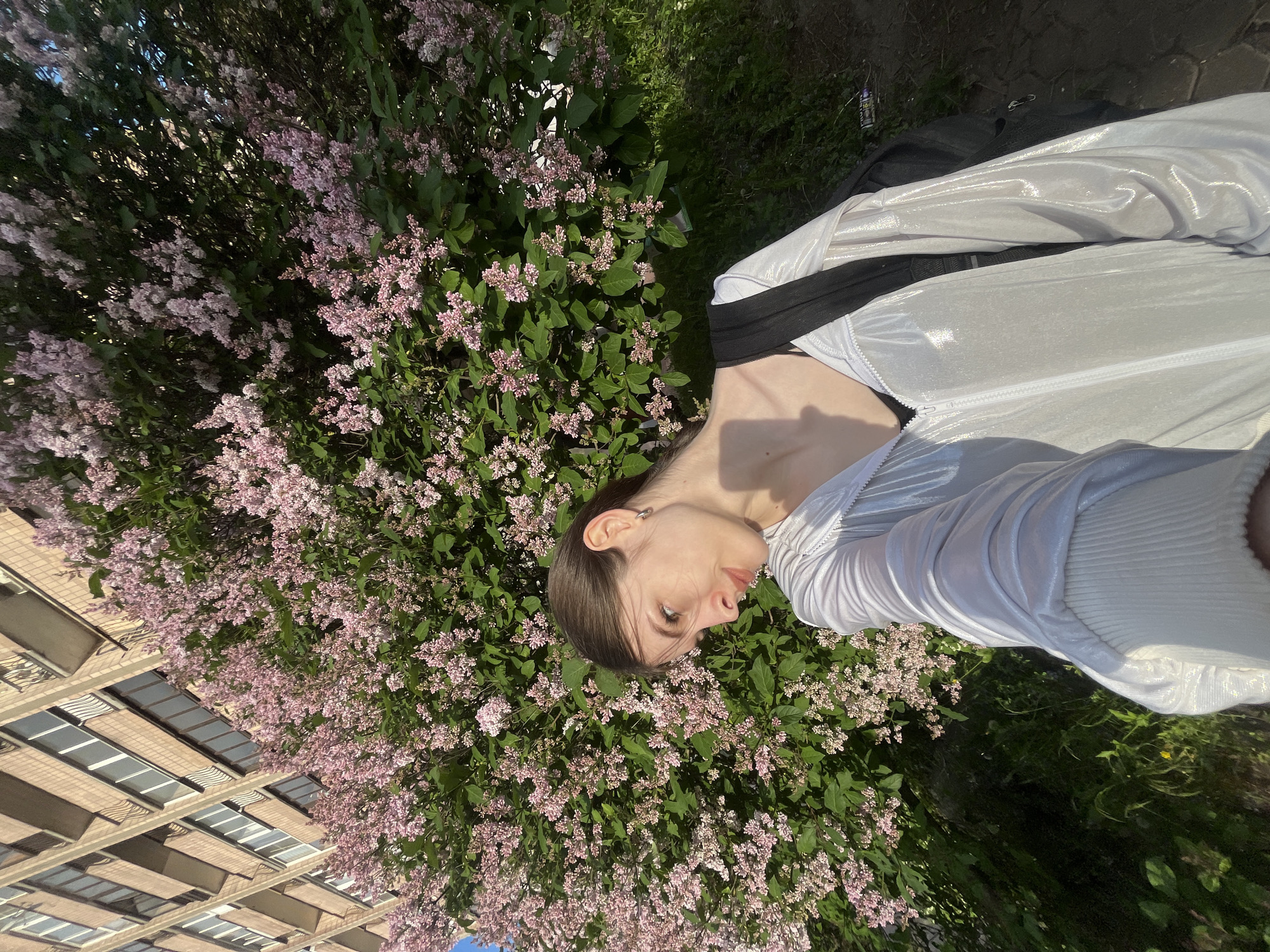 This bright, sunlit outdoor photograph, rotated 90 degrees clockwise, captures a young person reclining amidst a large lilac bush. The individual, with dark hair and a neutral expression, wears a shiny, light-colored jacket over a dark garment, and has a silver hoop earring. Their head is positioned towards the left side of the frame, looking slightly upwards.
The dominant feature of the scene is a lush lilac bush, filled with clusters of light pink and purple blossoms and green leaves, which spans the left and central portions of the image. Strong sunlight casts shadows, particularly visible on the person's neck and chest.
In the upper left background, part of a multi-story building with a light-colored facade and numerous windows is visible through the foliage. To the right of the person, patches of green grass and dirt are visible on the ground, along with a black backpack and a discarded metallic can.
The image conveys a sunny day, likely during spring or early summer, given the blooming lilac. The setting appears to be an urban or suburban environment, specifically in Saint Petersburg, Russia, featuring both natural elements and architecture. No visible text is present in the media.