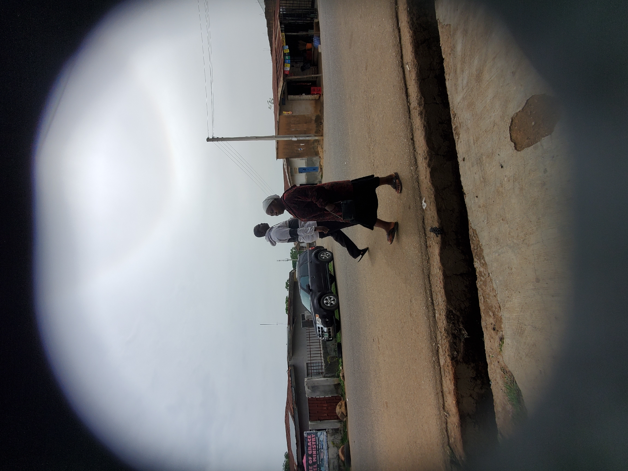 Two individuals are walking on a paved road in Jos, Nigeria. The first person is wearing a dark-colored top with a maroon long-sleeved shirt, a white head covering, and sandals. The second individual has on a white collared shirt with black pants. A dark-colored car is parked on the side of the road. Buildings line the street on either side. Utility poles and power lines are present. The sky is overcast. The image is viewed through an unknown circular aperture that causes dark vignetting. A concrete curb and drainage ditch run along the right side of the road.